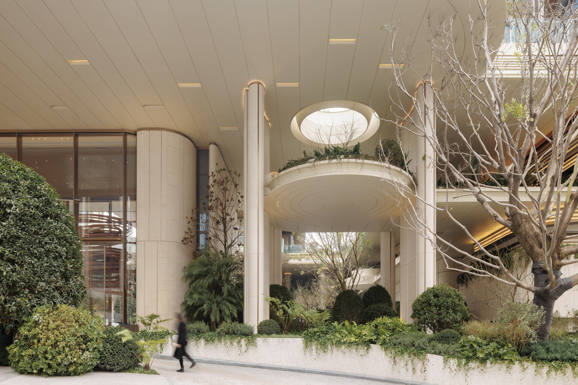 Round planter balcony with a skylight oculus above, between slender stone columns and a bare tree, as a pedestrian passes in soft daylight