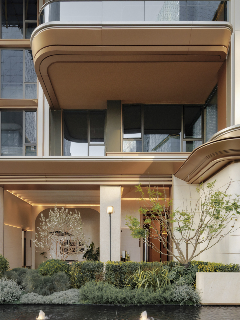 Bronze-toned curved balcony soffit above a glazed entrance, with clipped hedges and a reflecting pool with small fountains in front