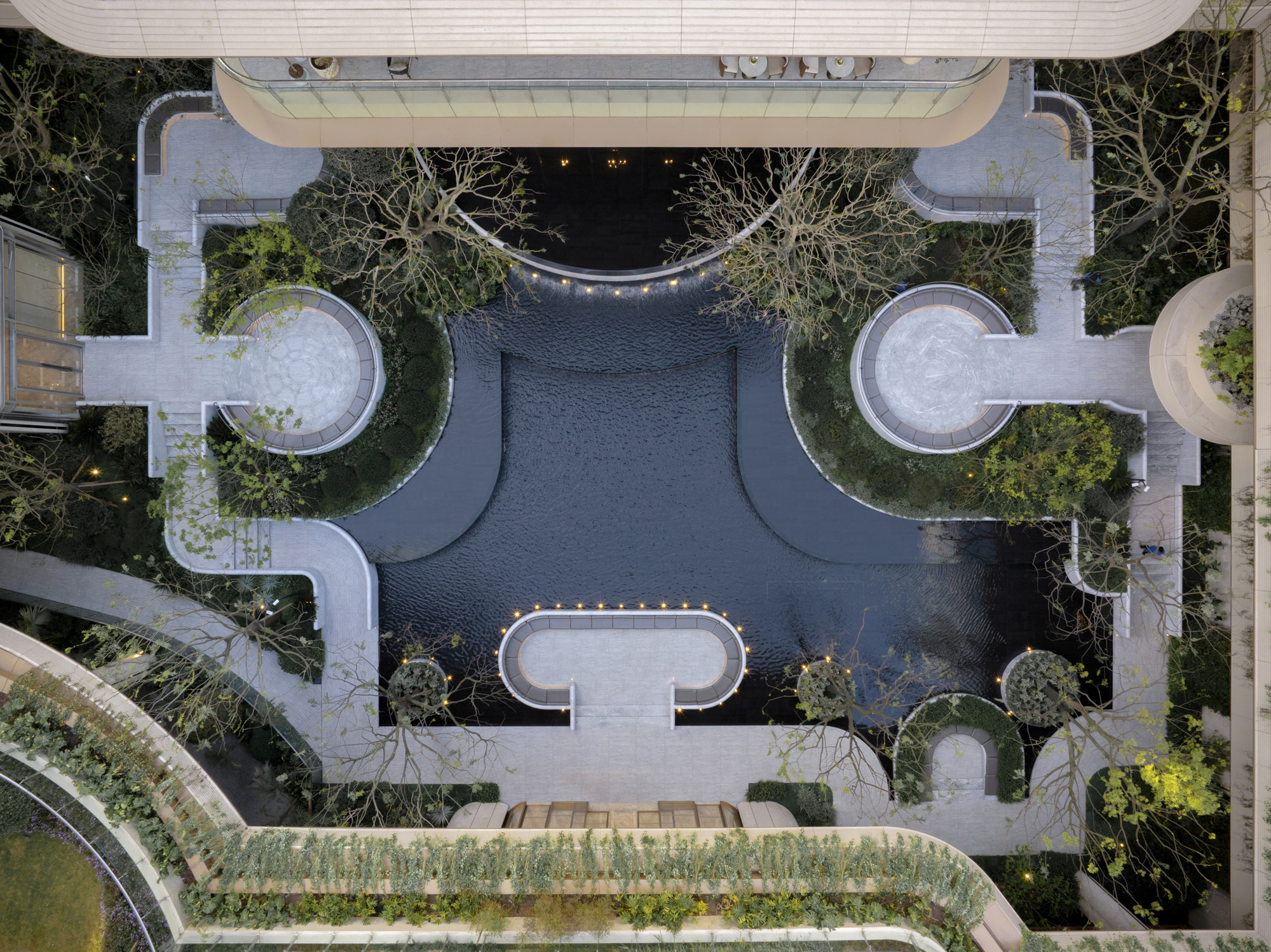 Top-down view of a dark reflecting pool with round stone seating platforms, curved paths and bare trees lit by small lights at dusk