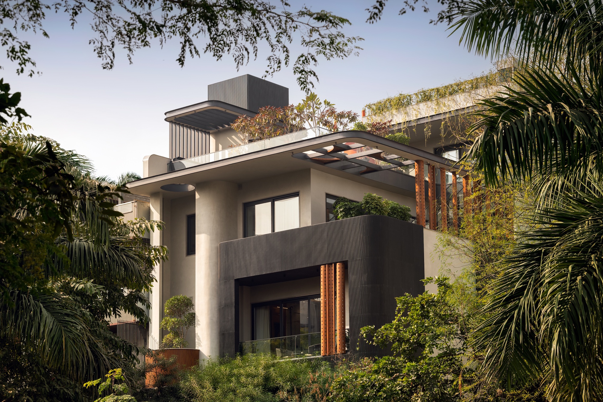 Upper floors of a stepped house with a curved canopy and terracotta screens, framed by palm fronds in clear daylight