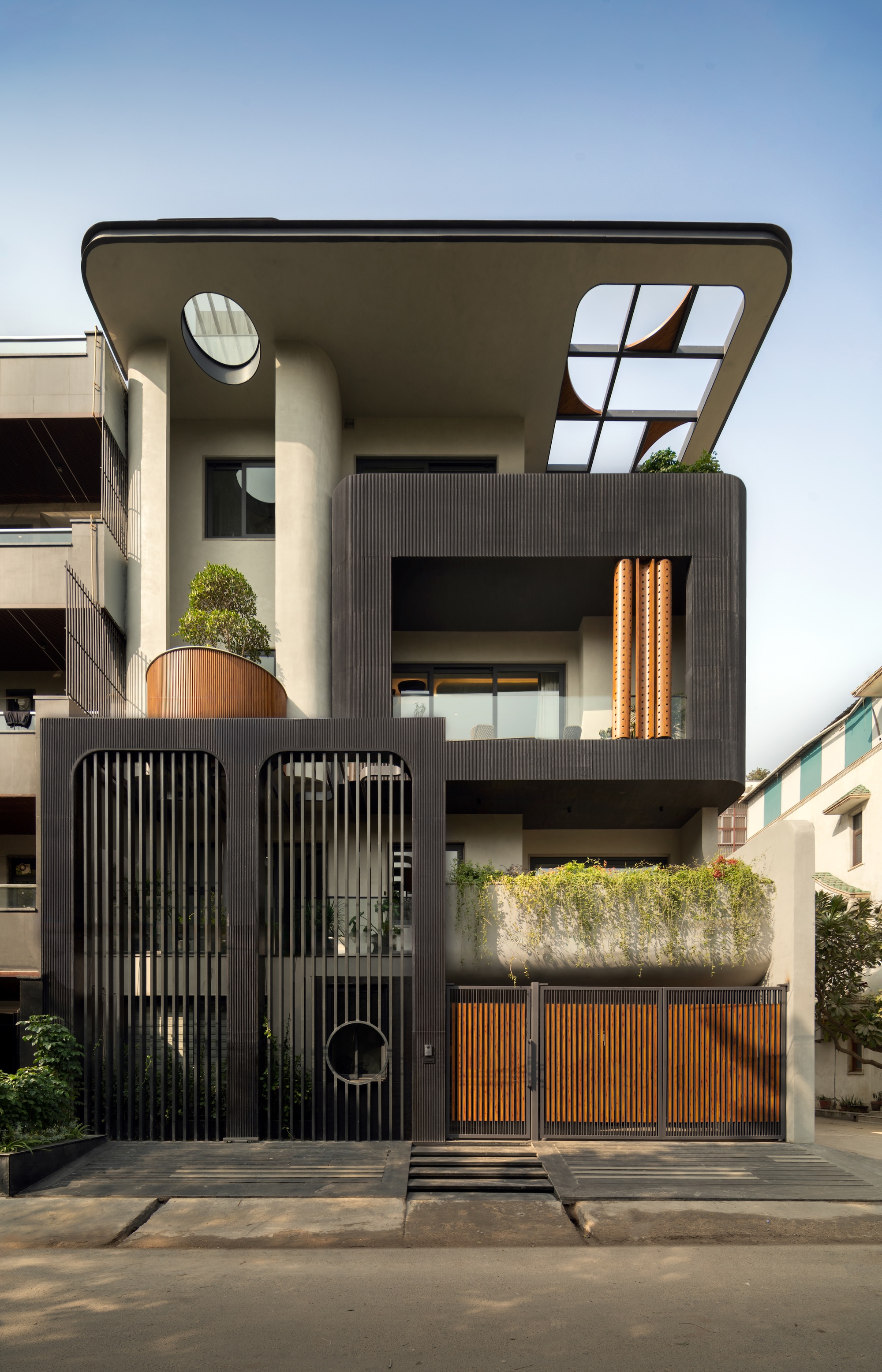 Street facade with a curved overhanging roof, oval skylight, slatted black gate and trailing planter, under a pale blue sky