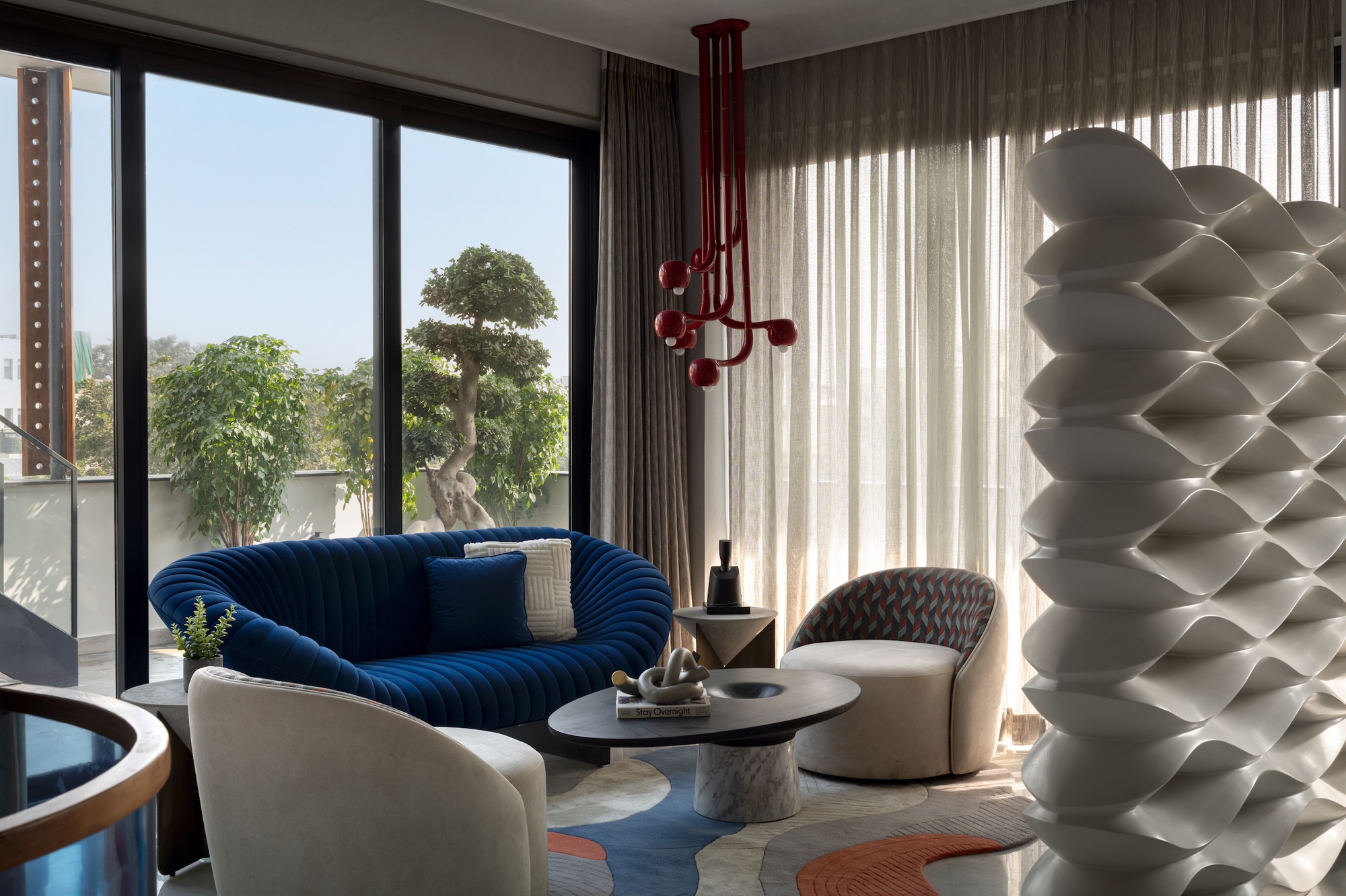 Living corner with a blue curved sofa, red pendant light and a white wave-patterned sculptural screen beside sheer curtains
