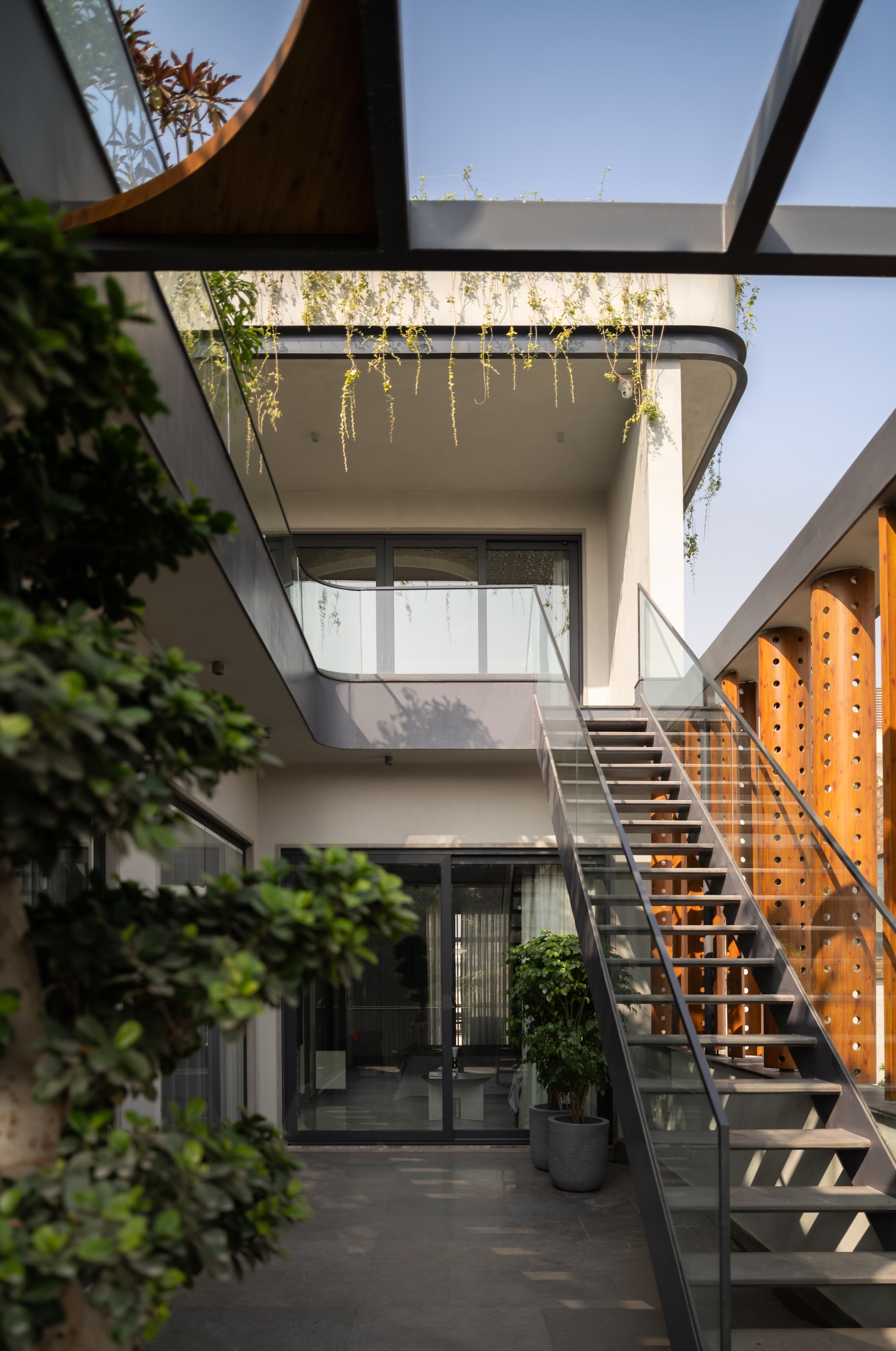 Steel staircase beside perforated timber columns, with a curved glass balcony and trailing vines overhead in clear daylight