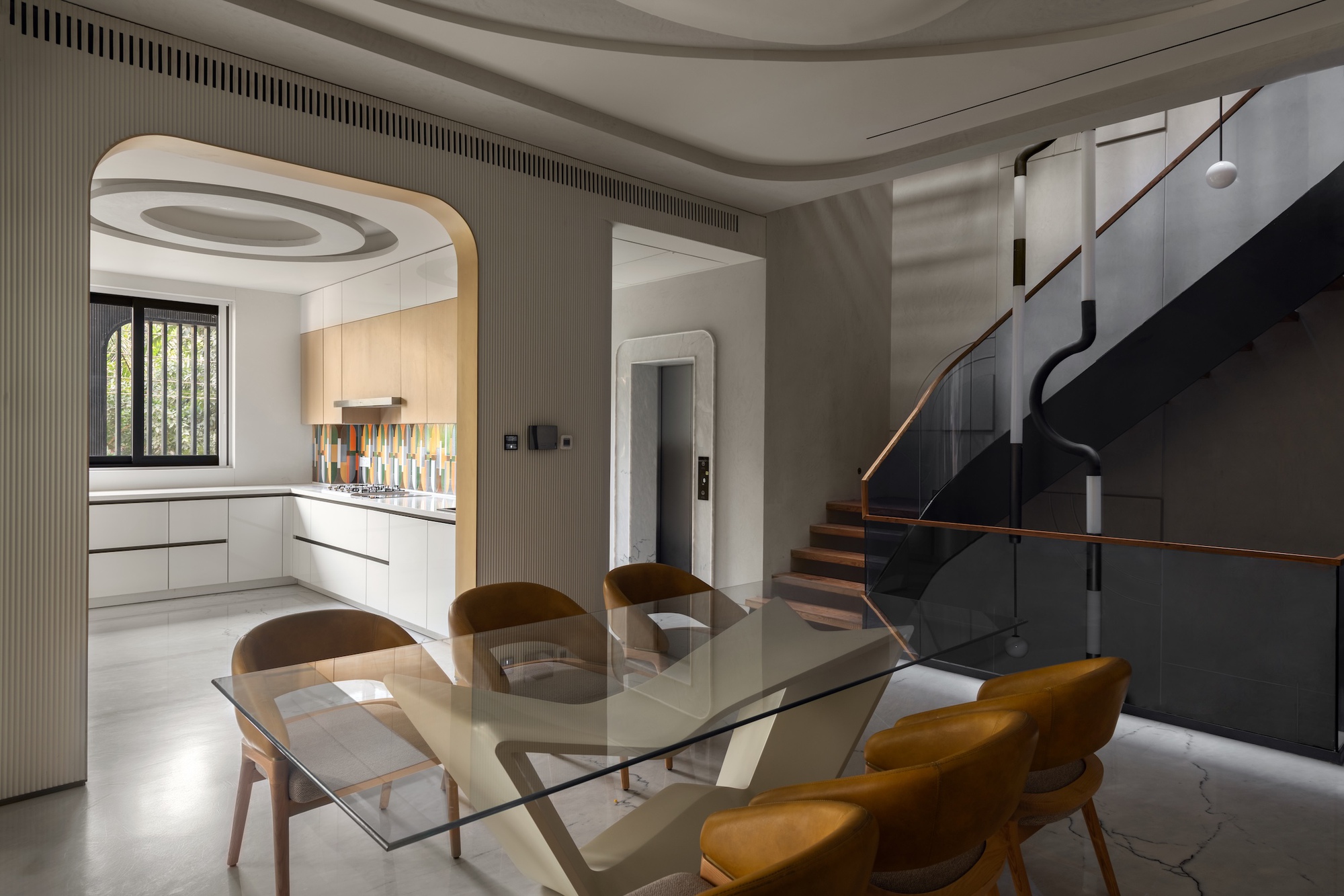 Glass-topped dining table with tan chairs in front of an arched kitchen opening and a black steel staircase