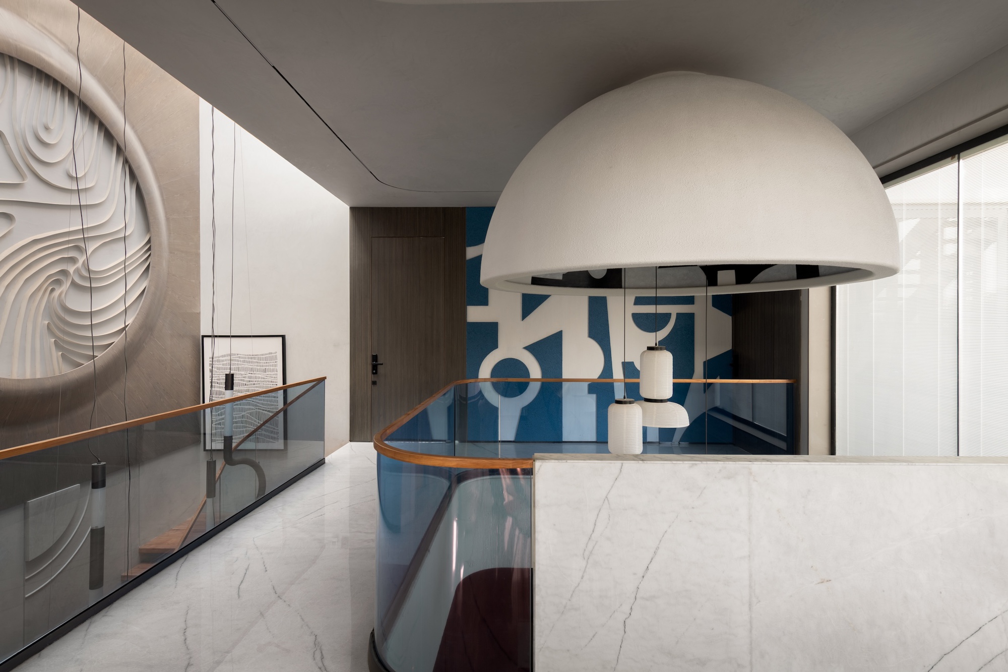 Upper landing with a large white dome pendant, curved glass railing, marble parapet and a blue geometric mural behind