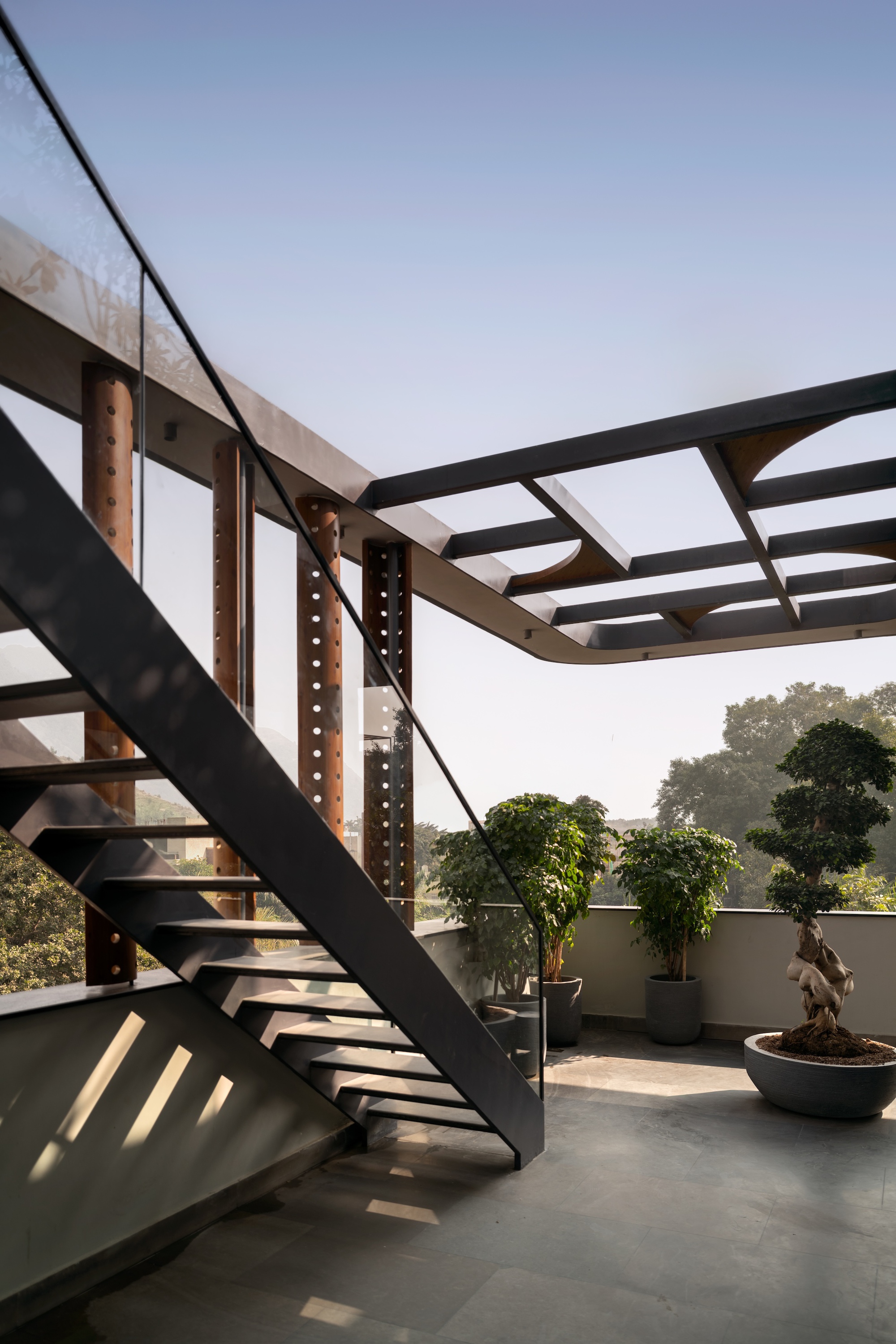Rooftop terrace with a steel stair, perforated rust-toned columns, a pergola frame and potted bonsai in low afternoon light