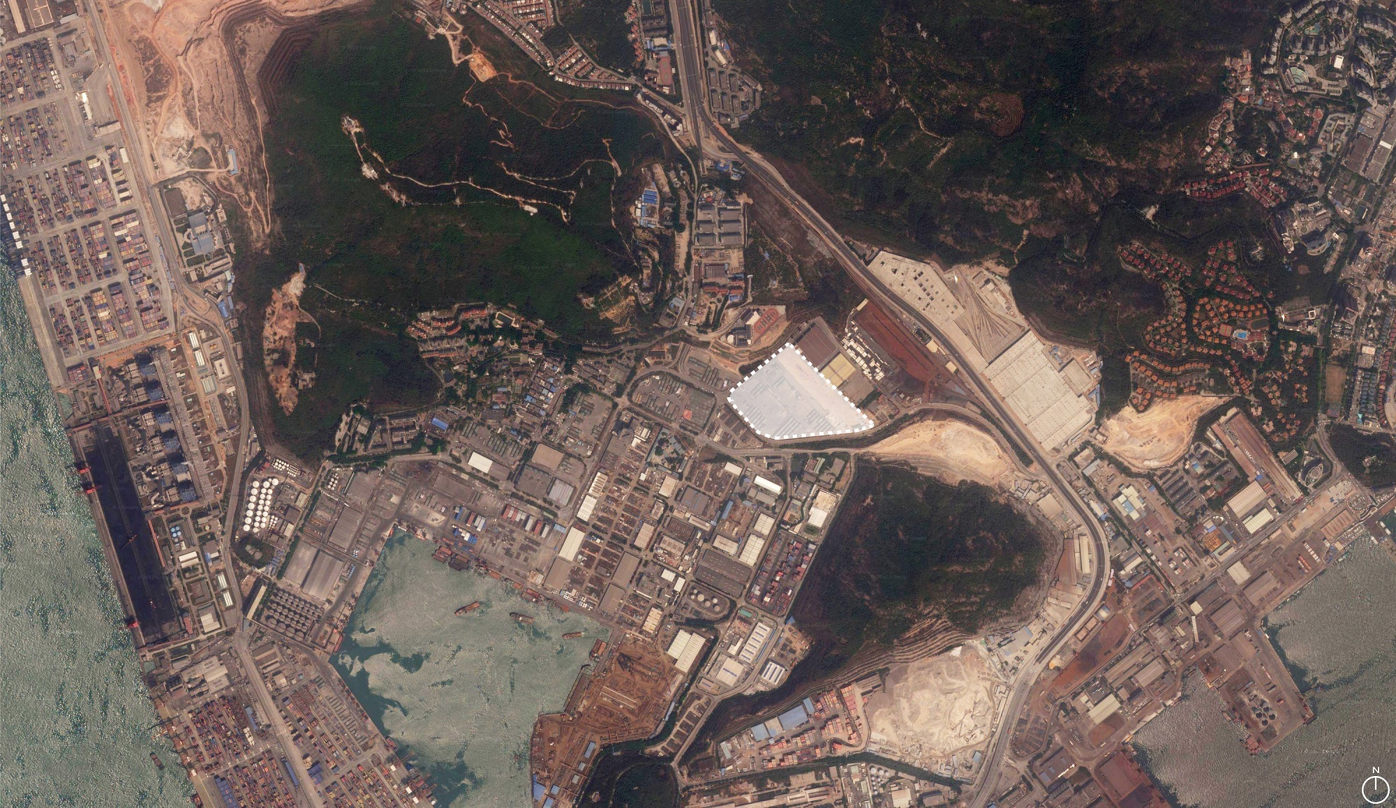 Aerial satellite view of a white-roofed building among container terminals, hillsides and industrial blocks beside a hazy bay