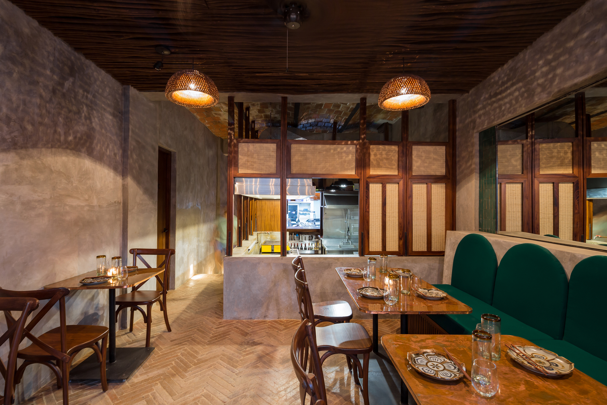 Dining room with green banquette, copper-topped tables and woven pendant lamps facing an open kitchen screened by timber and cane panels