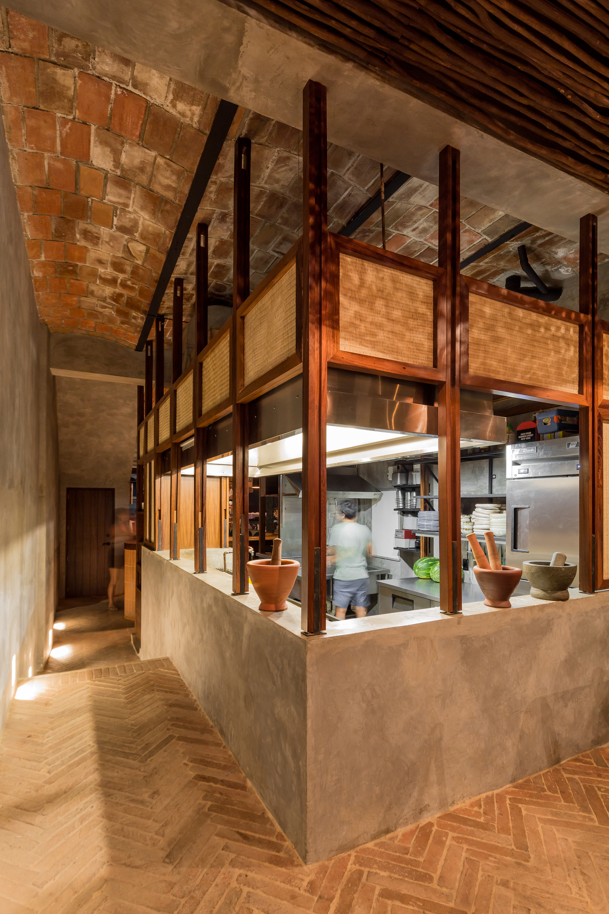 Open kitchen counter in a plastered block with timber posts and cane panels, a cook working beneath a brick vault