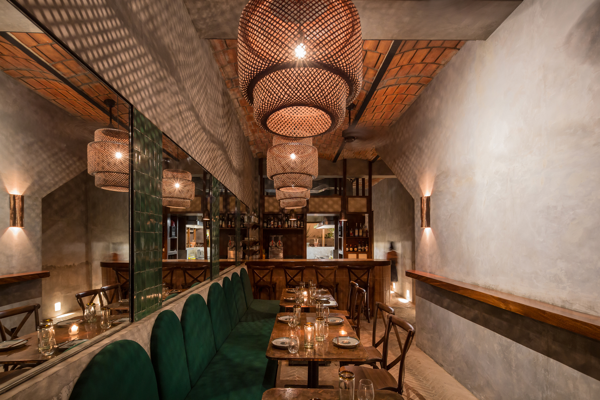 Long dining room with green tile banquette, mirrored wall and tall woven pendants glowing in the evening, bar at the far end