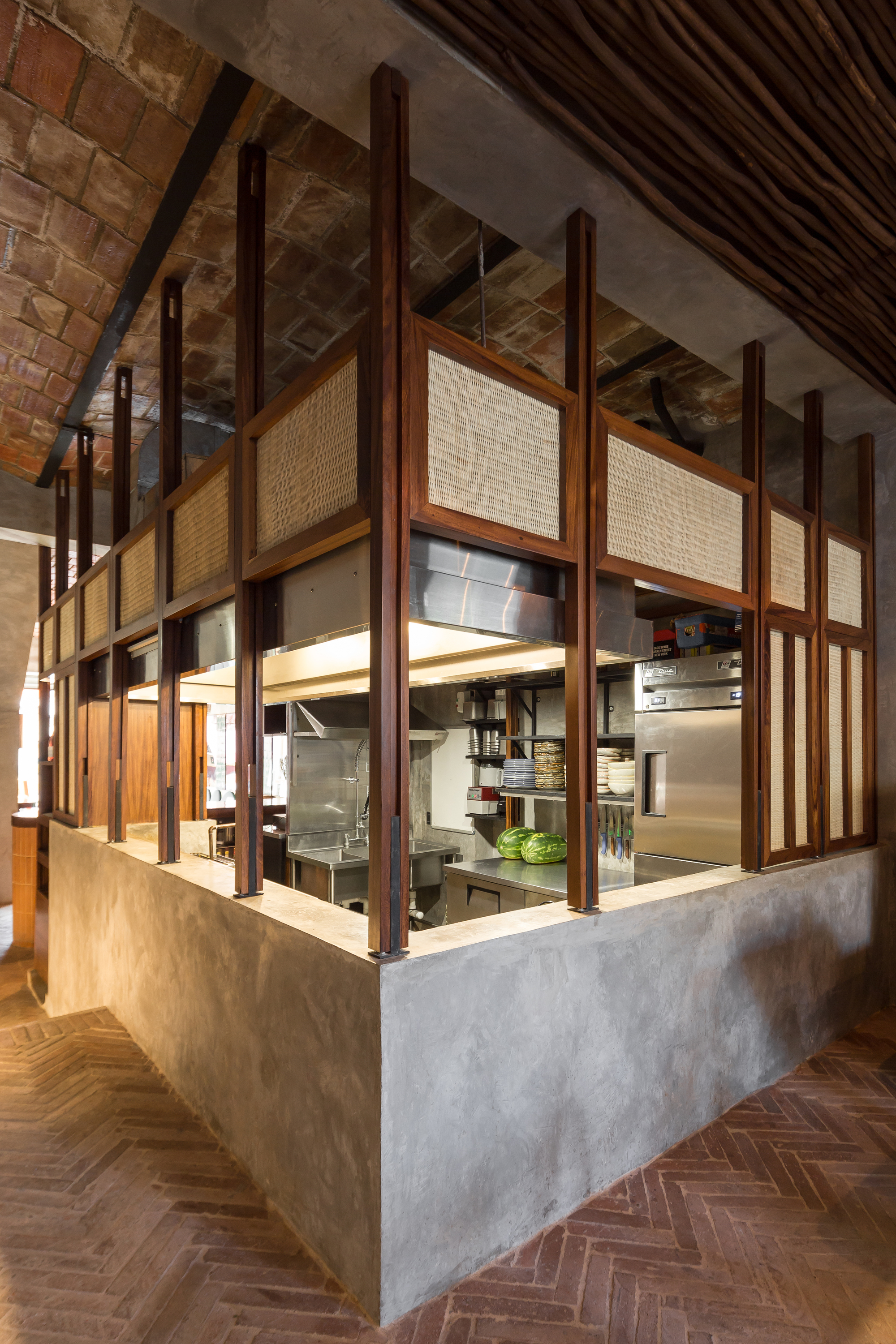 Corner of the kitchen enclosure with dark timber posts, cane infill panels and watermelons on the steel counter below a brick vault