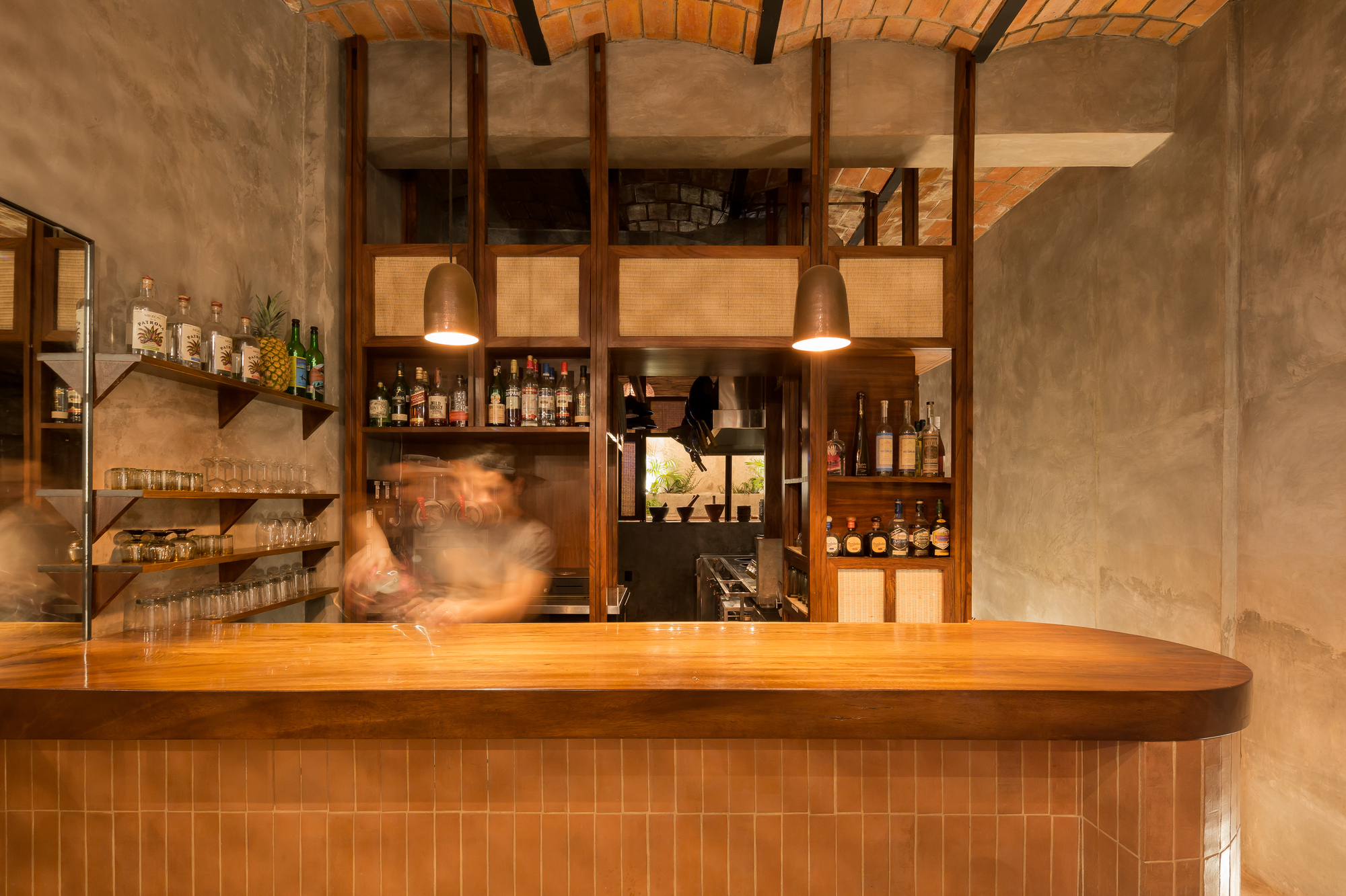 Curved timber bar top over orange tile base, with a bartender blurred in motion before a rattan-panelled bottle shelf under a brick vault