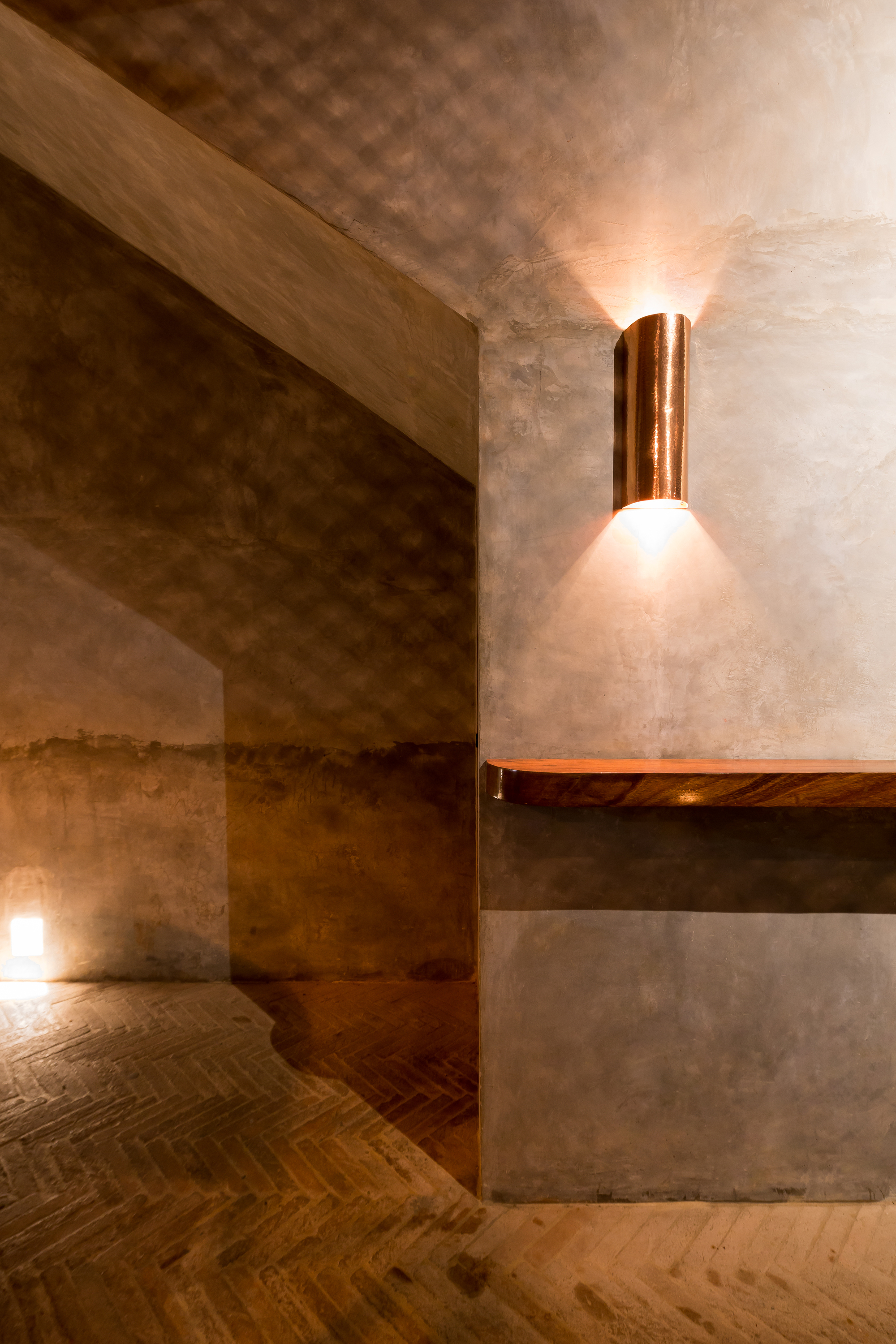 Copper wall sconce casting a fan of light across plastered walls above a herringbone brick floor