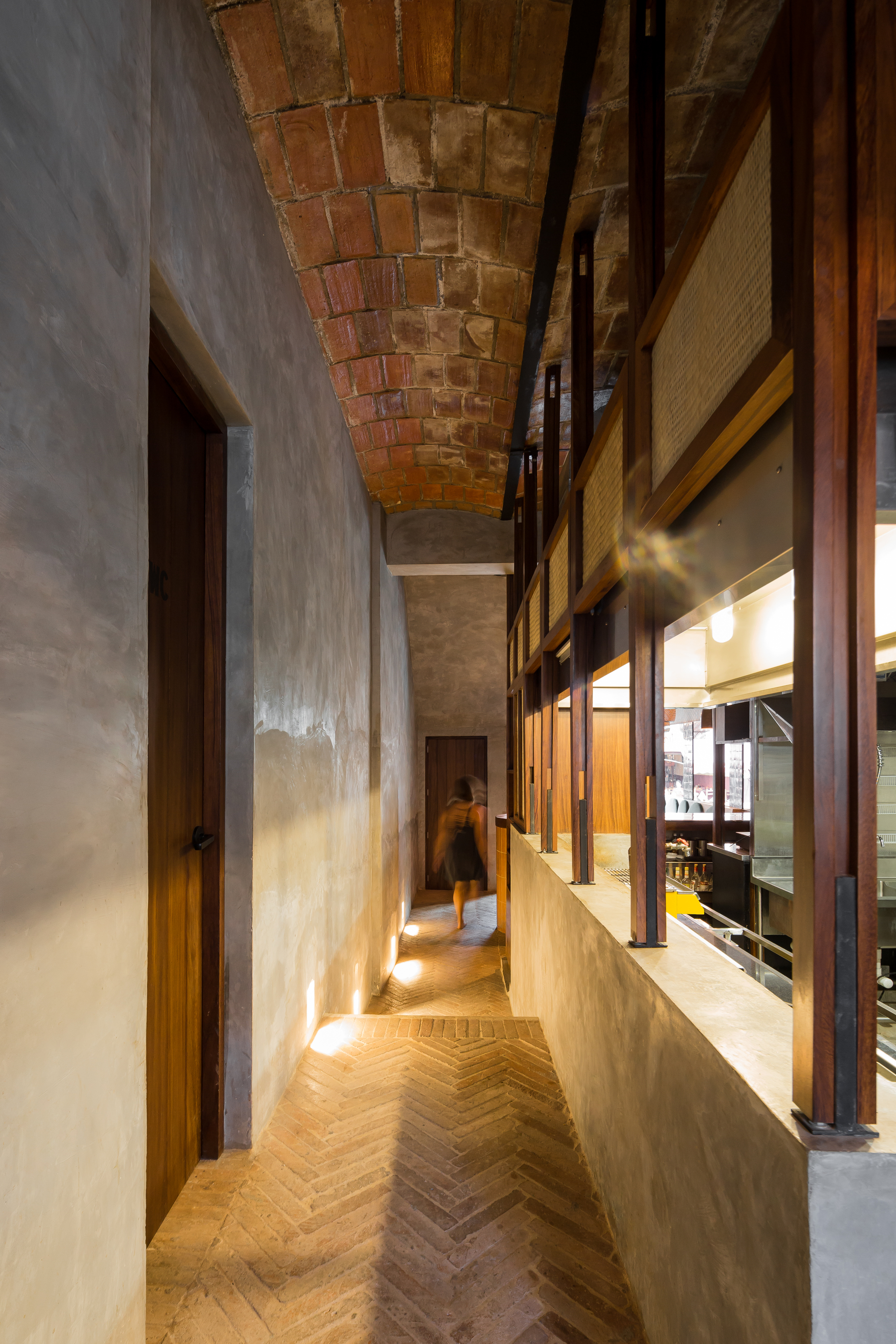 Narrow herringbone brick corridor under a brick barrel vault, with a woman walking past a timber-framed kitchen pass