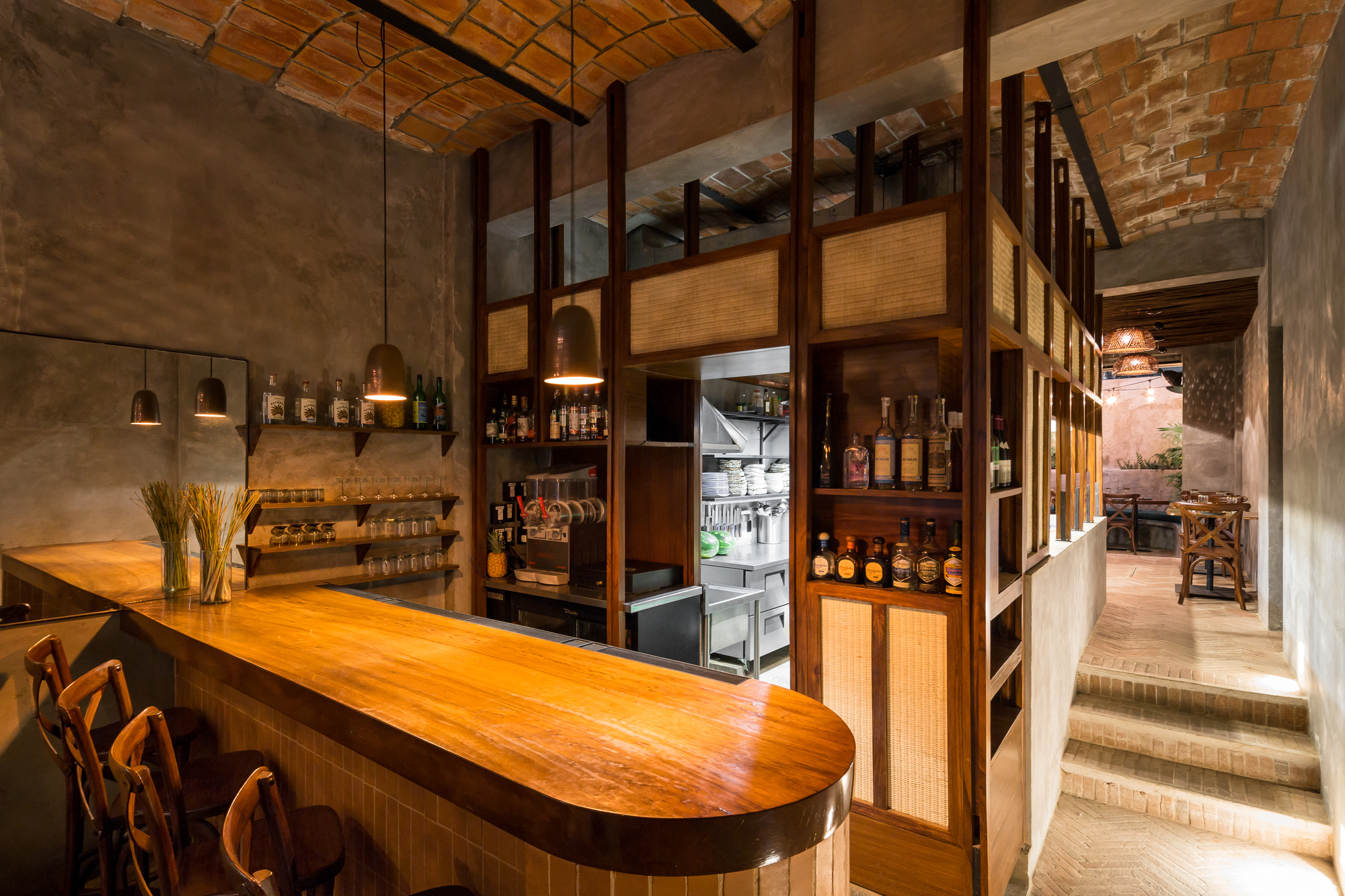 Rounded timber bar counter facing a wood and rattan back shelf, with brick steps rising toward the dining area at right