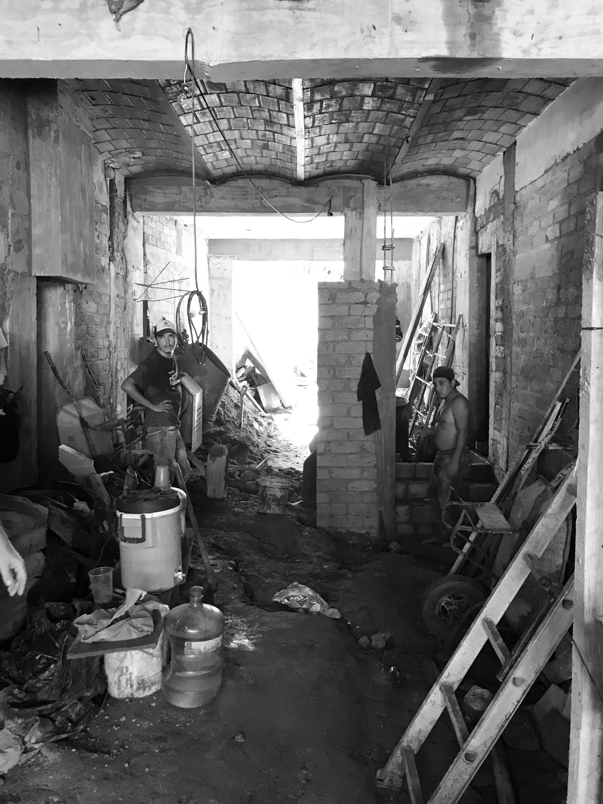 Black and white construction site view of a narrow brick-walled passage with a vaulted brick ceiling and two workers amid ladders and debris