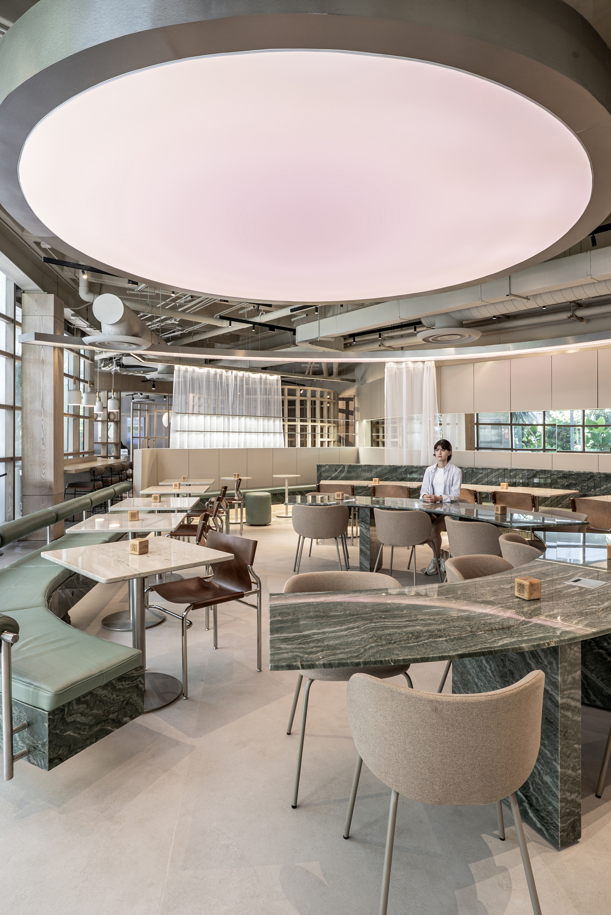 Vertical view of the pink luminous ceiling disc over marble-edged tables, a green banquette and a woman seated beyond