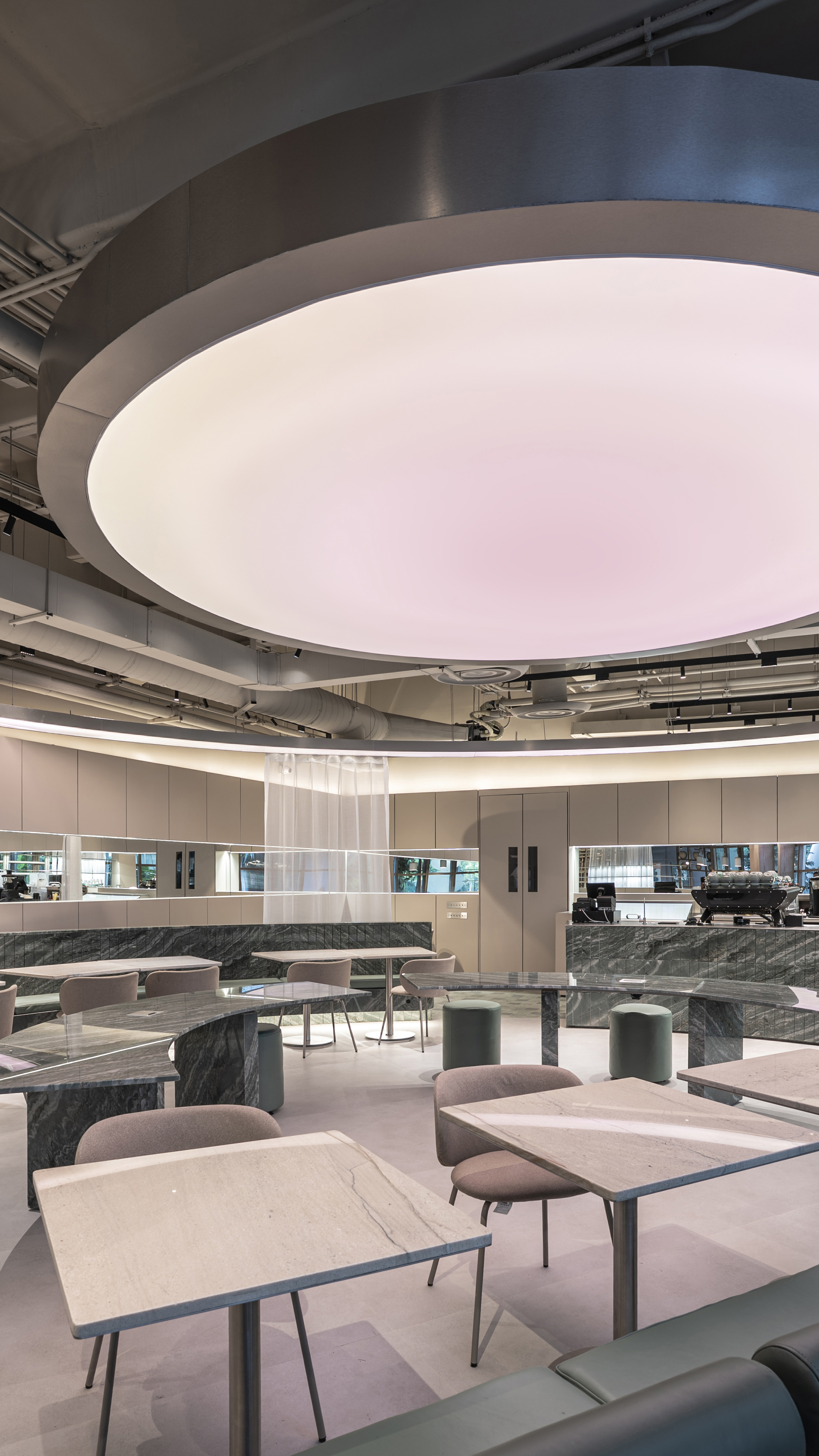 Looking up at the pink glowing ceiling ring above square stone tables, green stools and a marble coffee bar at the back