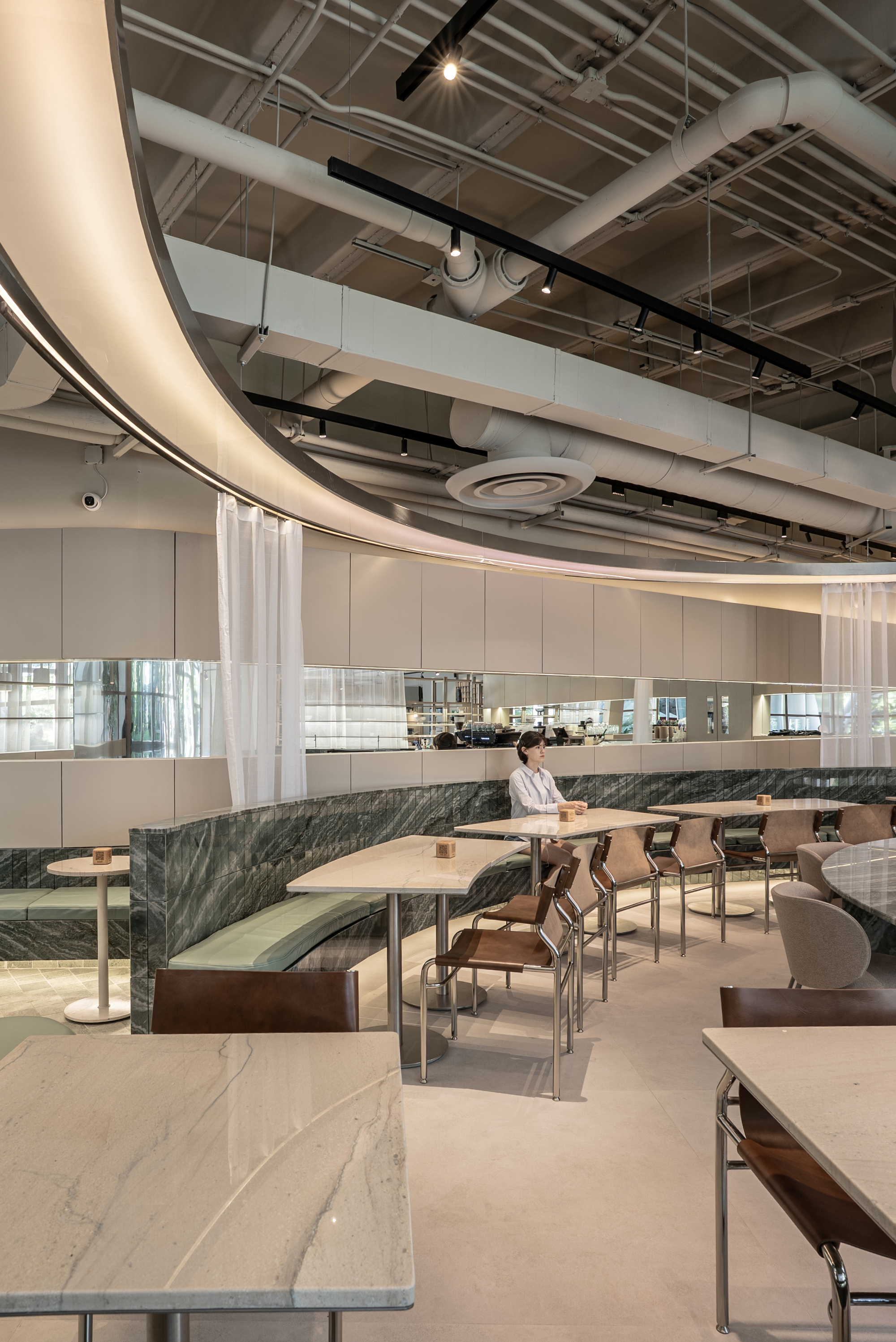 Sweeping curved soffit with exposed white ducts above a marble banquette, leather chairs and a woman seated at a table