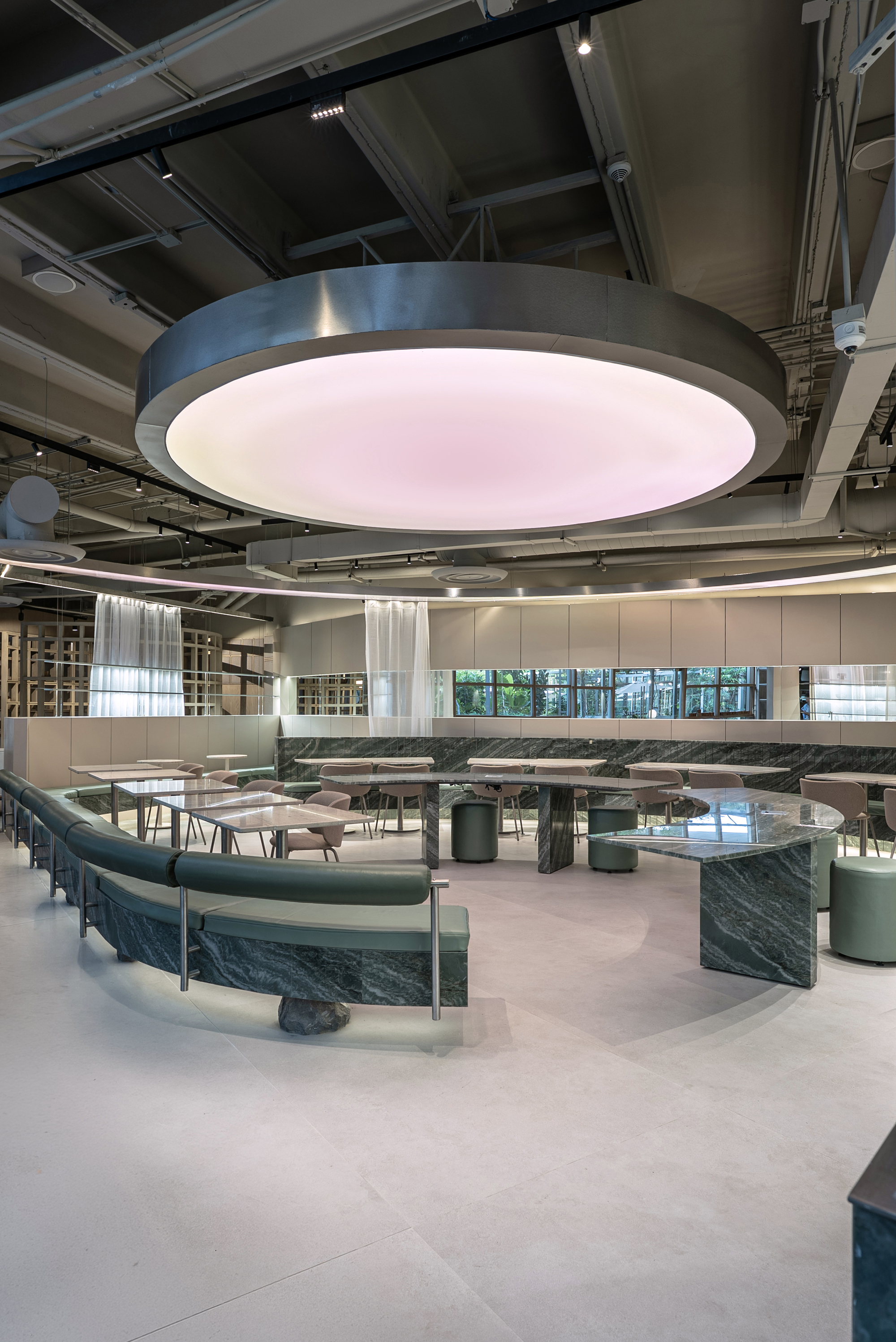 Curved green banquette and veined stone table beneath a round glowing pink ceiling disc with a steel rim