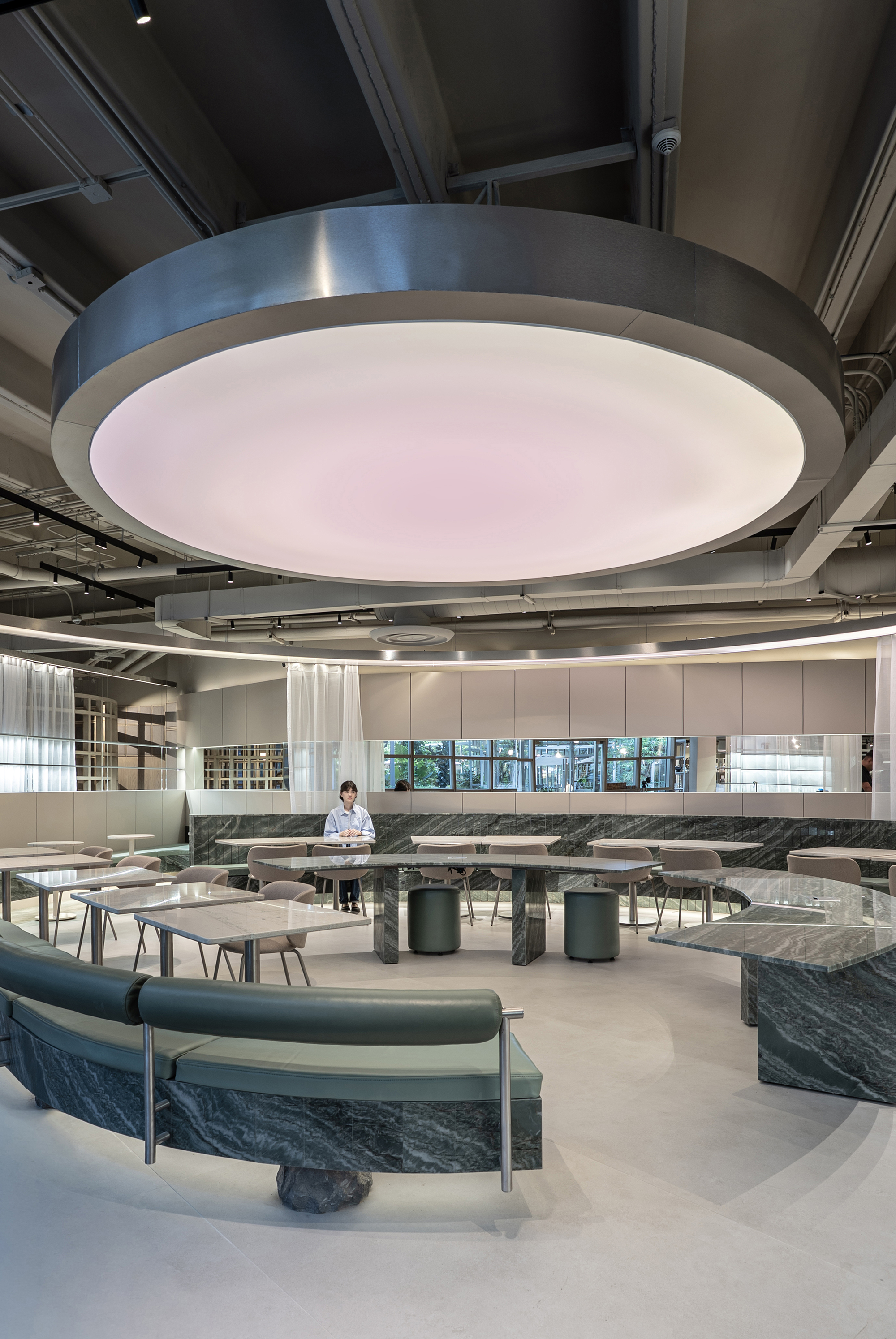 Circular backlit ceiling canopy above stone-based tables, with a lone diner seated near the garden-facing windows