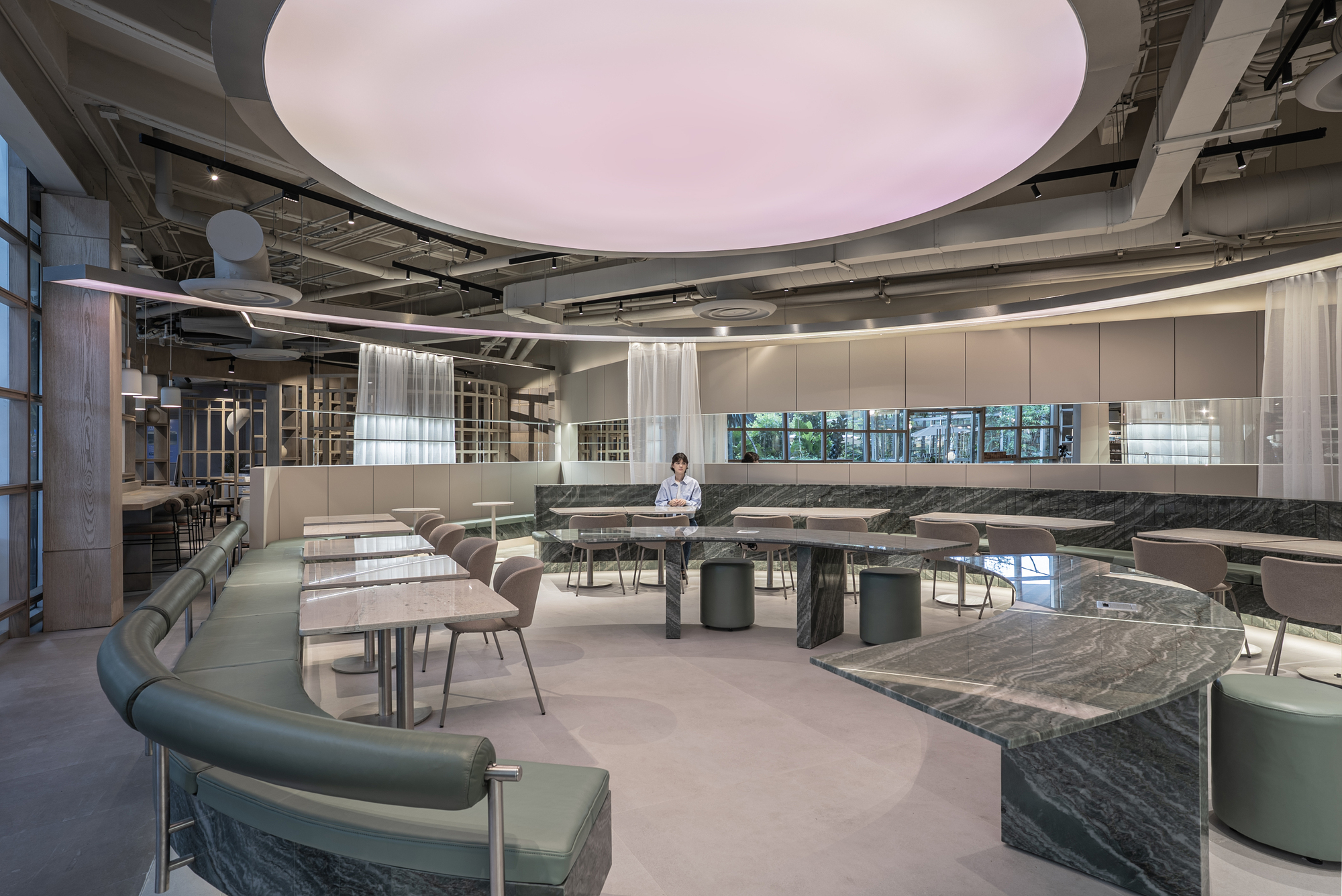 Wide view of the dining hall with curved banquette, stone tables and a seated visitor under the glowing ceiling disc