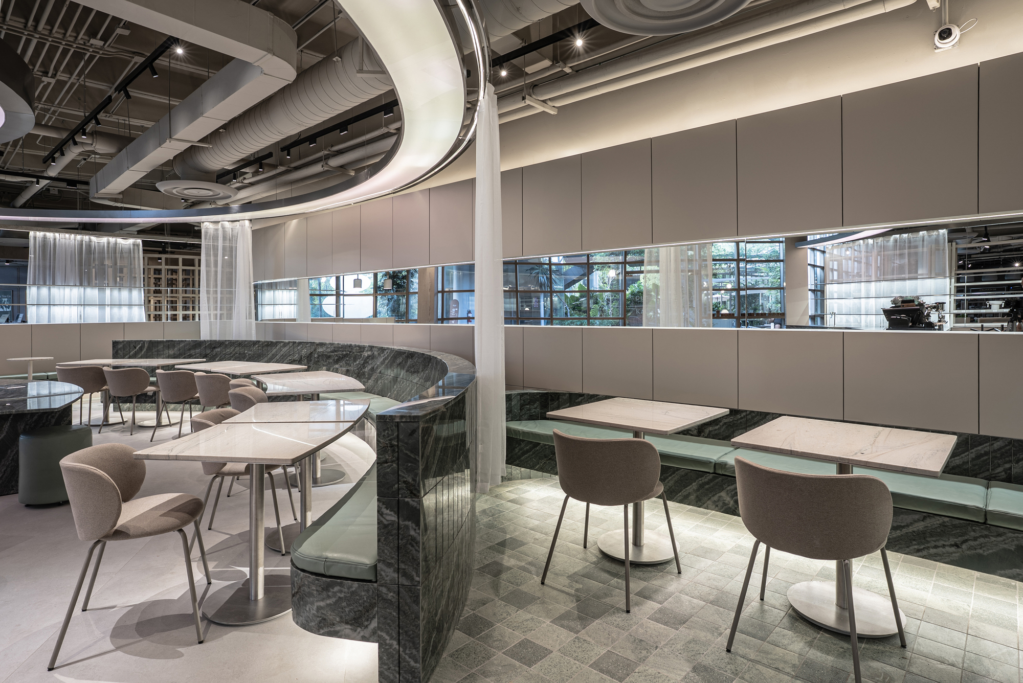 Curved green marble banquette edge beside square tables and taupe chairs, with a mirrored band reflecting garden windows