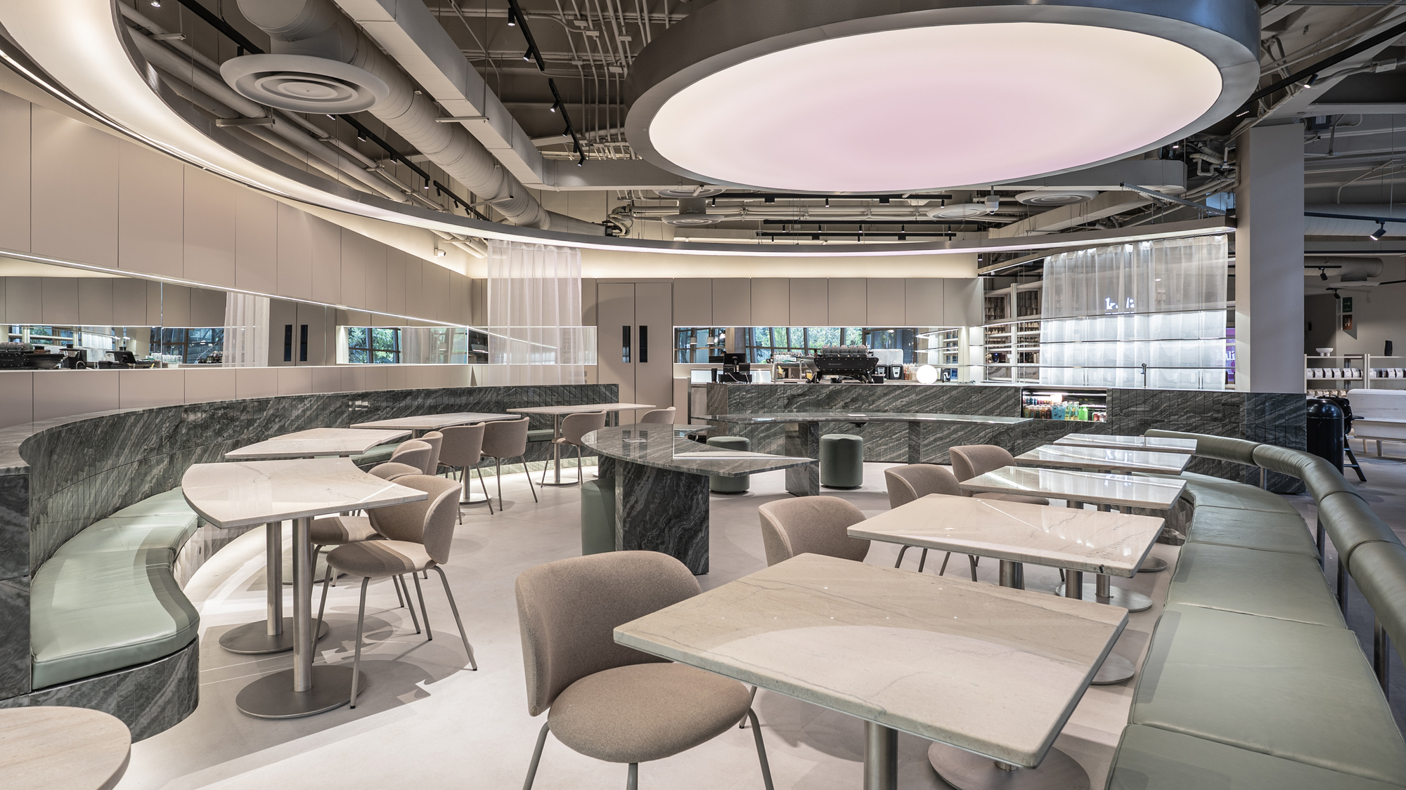 Wide view of the dining hall with a glowing pink oval ceiling light above marble tables and sage banquettes