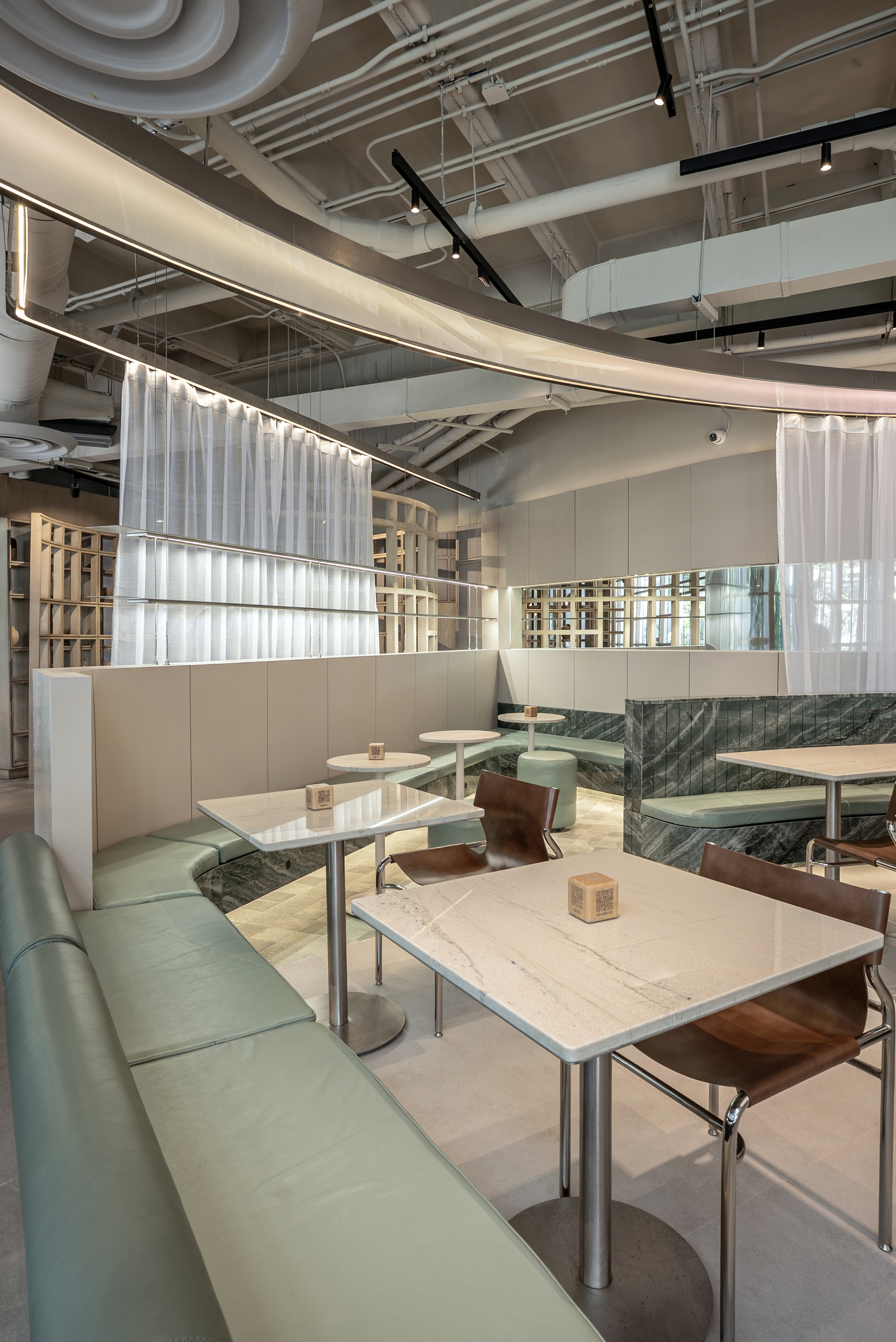 Sage leather banquette curving past small marble tables and brown leather chairs, with sheer curtains lit from behind