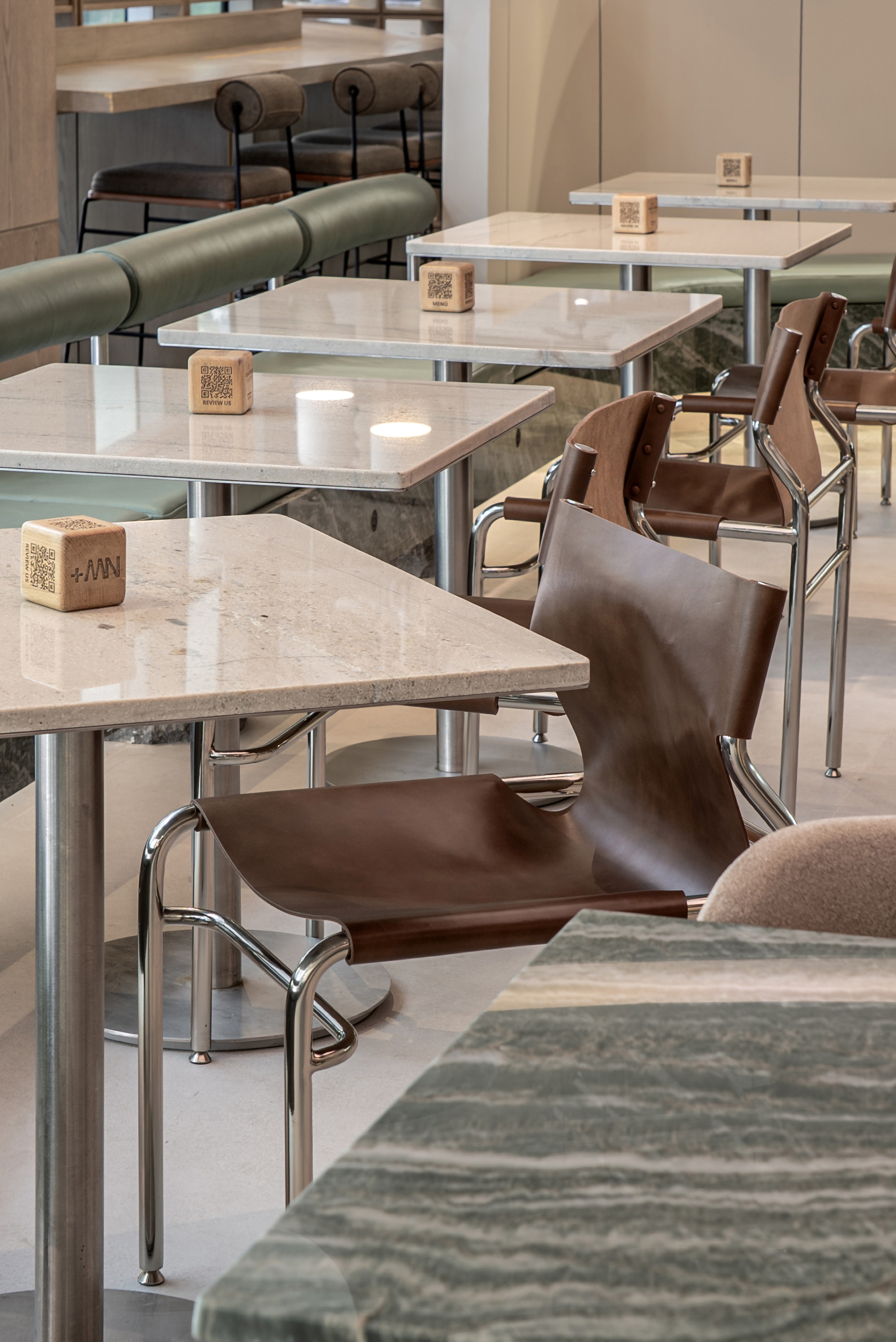 Row of square stone-topped tables with brown leather sling chairs, steel frames and small wooden cubes under soft daylight
