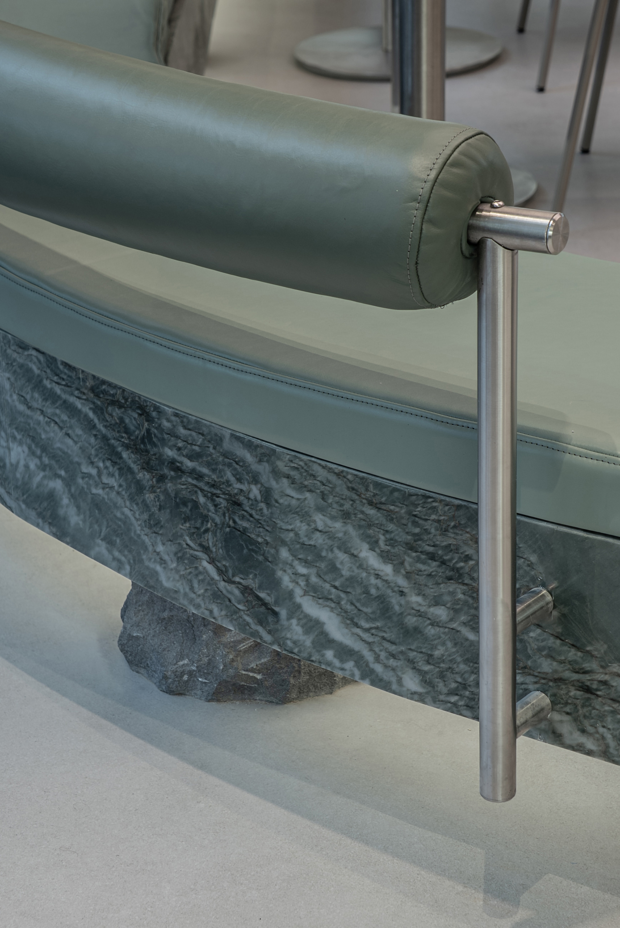 Close-up of a curved bench with green leather bolster, rough-cut stone base and brushed steel upright on a pale floor