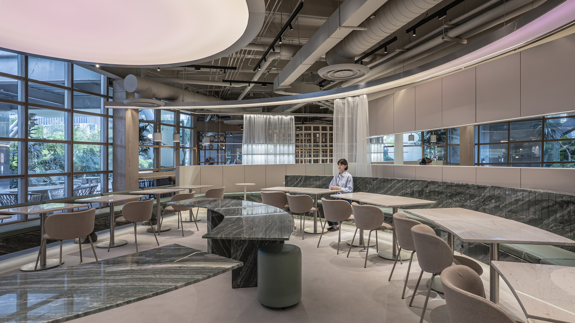 Dining hall with curved stone tables, pale chairs and a seated woman beside a glazed wall overlooking planting