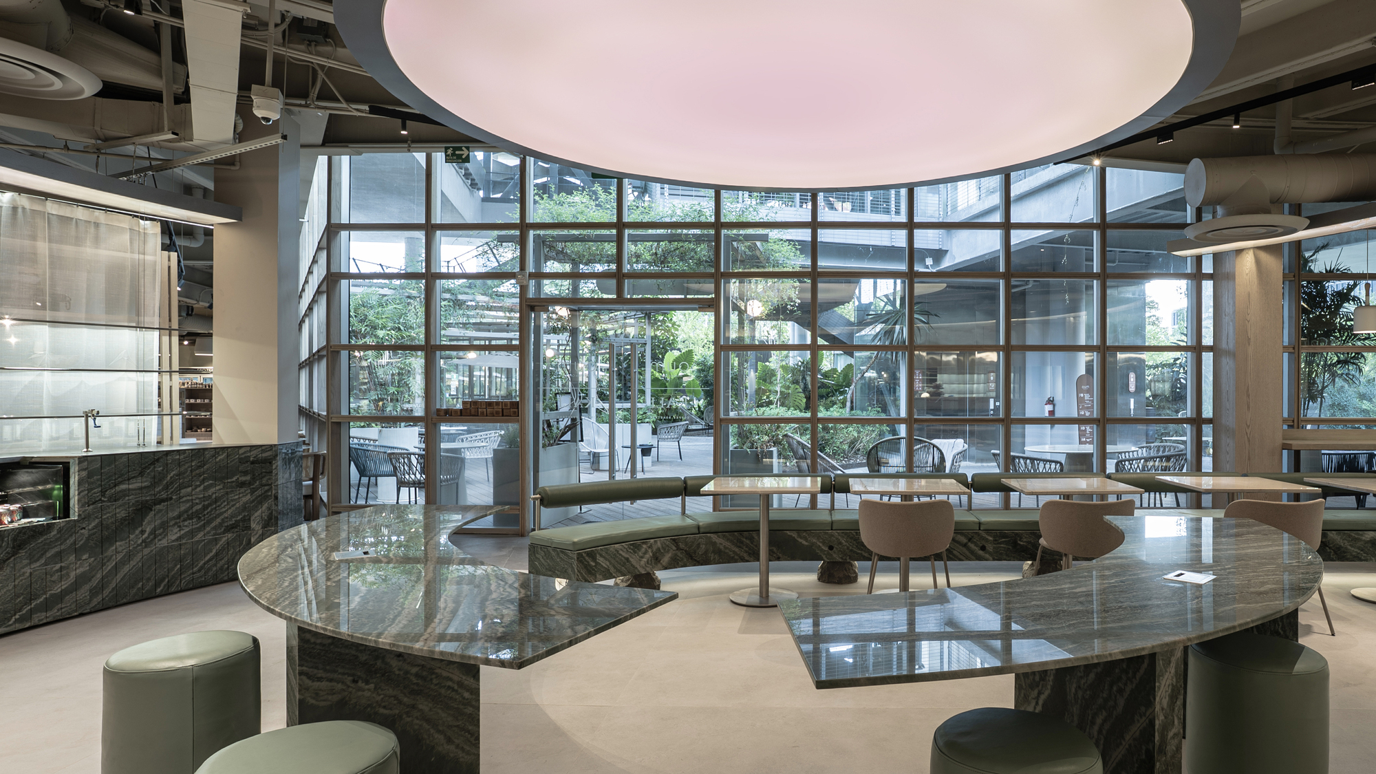 Round stone tables and green leather stools under a glowing pink ceiling disc, facing a gridded glass wall and garden