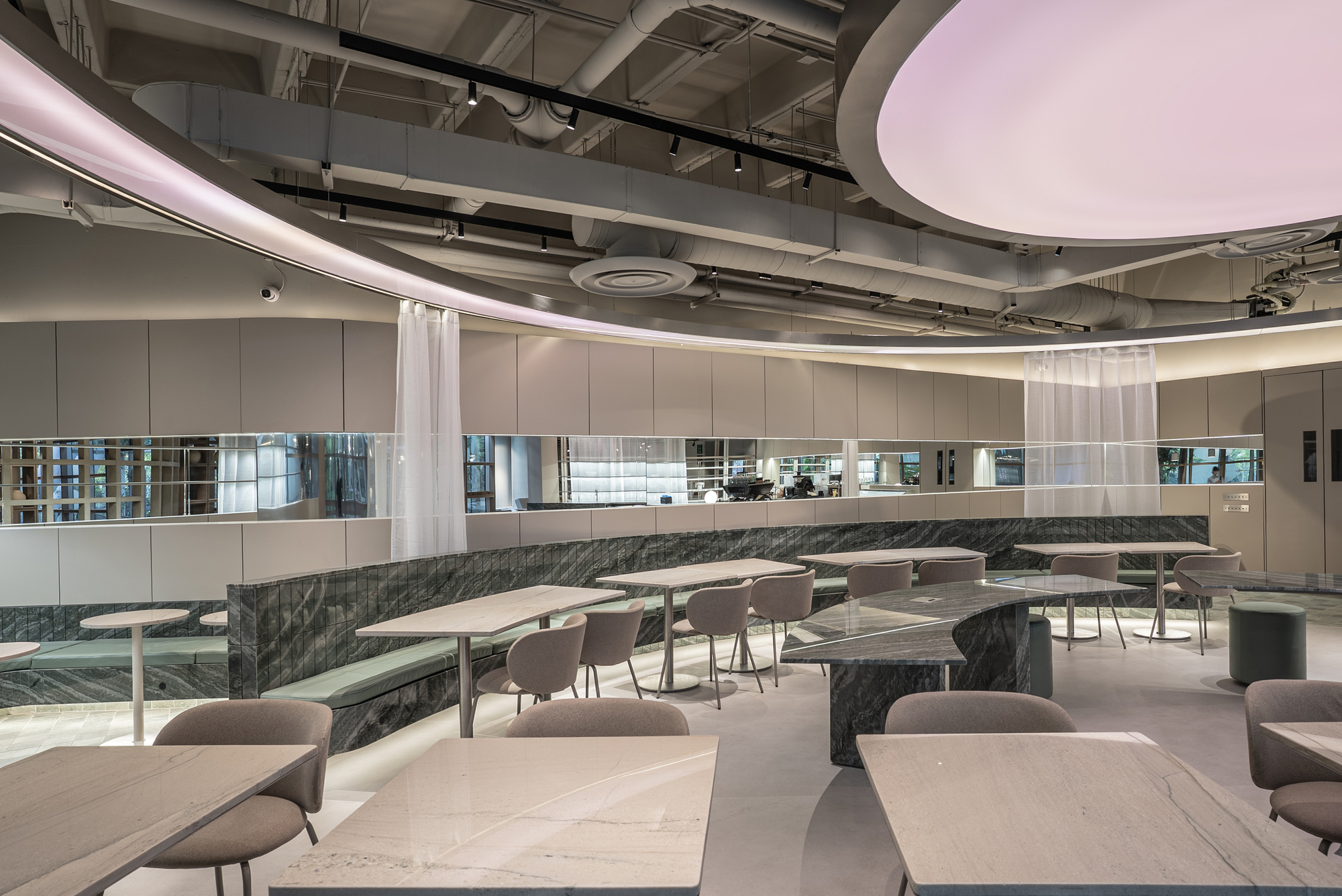 Curving wall of panels and mirror strips above a stone-clad banquette, with sheer curtains and a lit ceiling ring over the tables