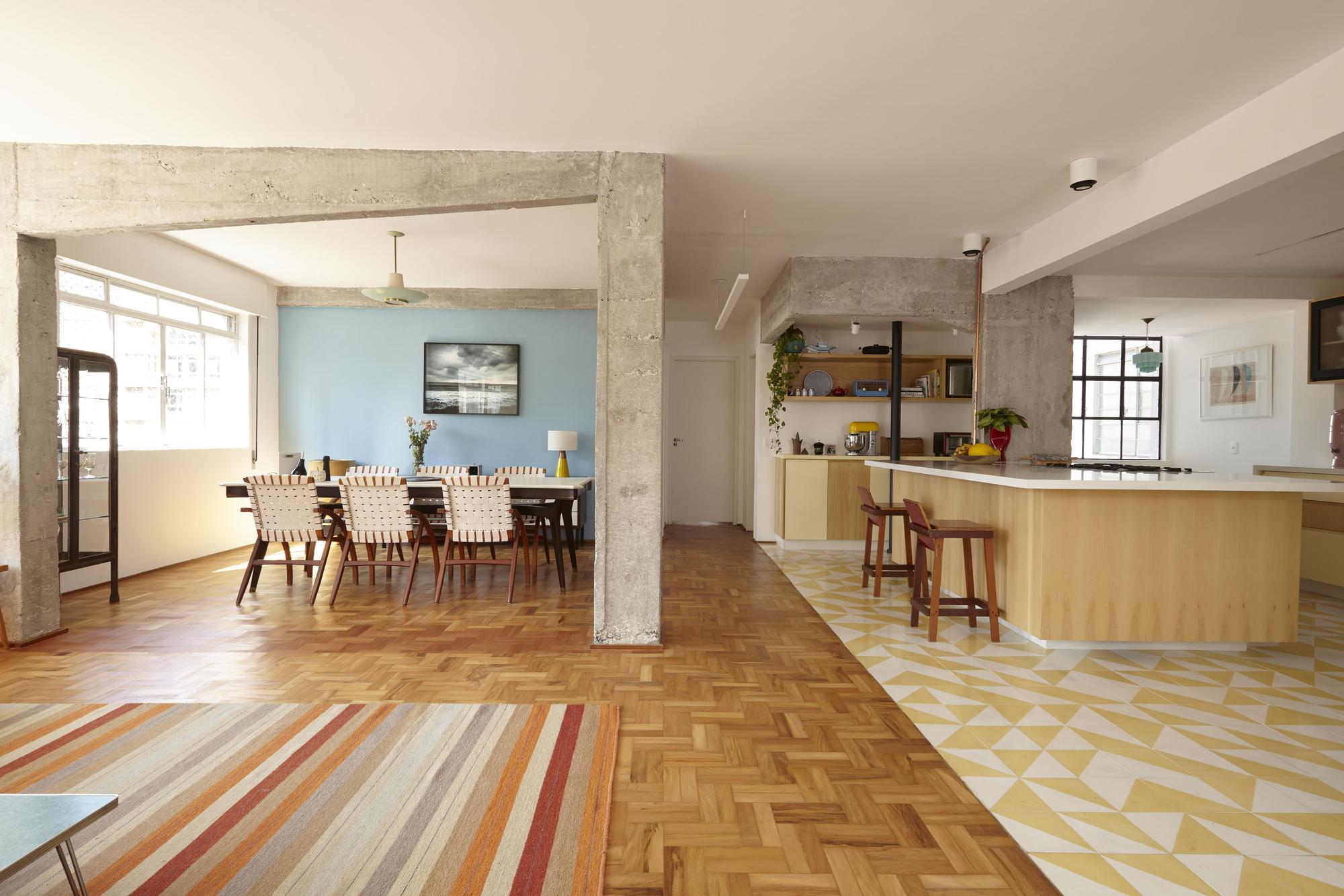Exposed concrete beam and column dividing a parquet-floored dining area from a kitchen with yellow geometric tile, in afternoon daylight