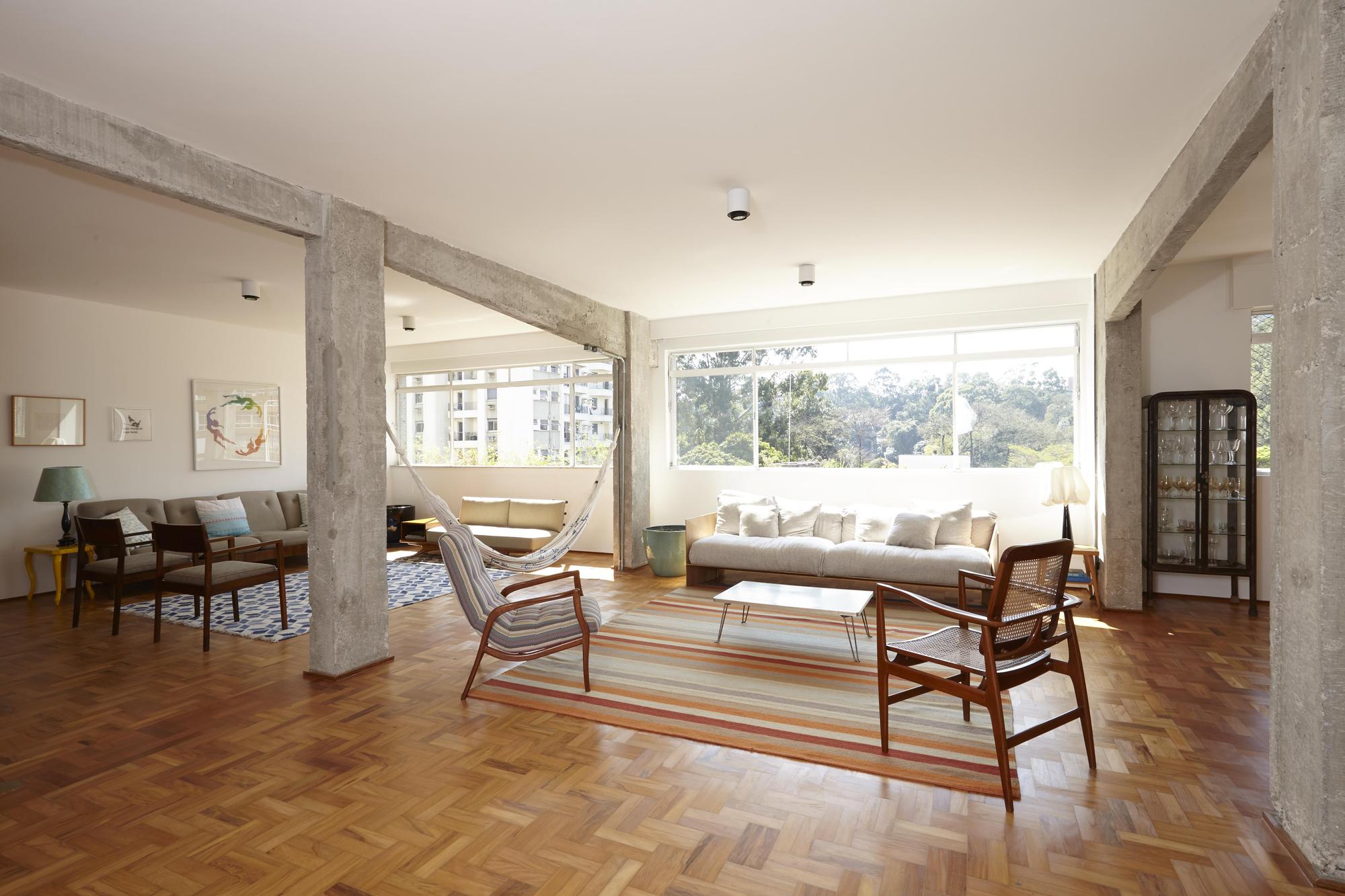 Living room with rough concrete columns, a hammock, cane armchairs and a striped rug facing tall windows onto treetops