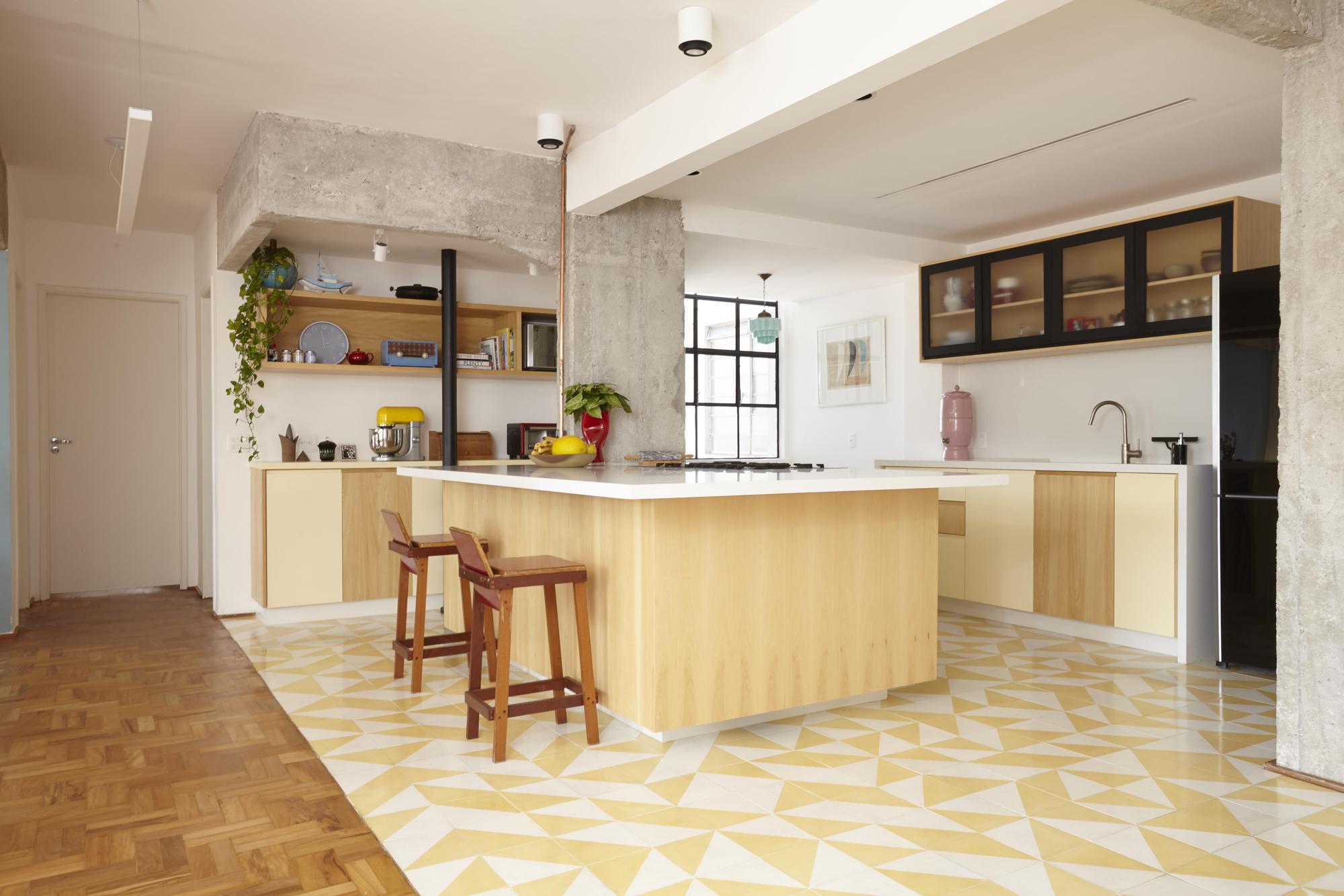 Kitchen island with pale wood cabinetry and red stools on a yellow geometric tile floor beside bare concrete columns in daylight