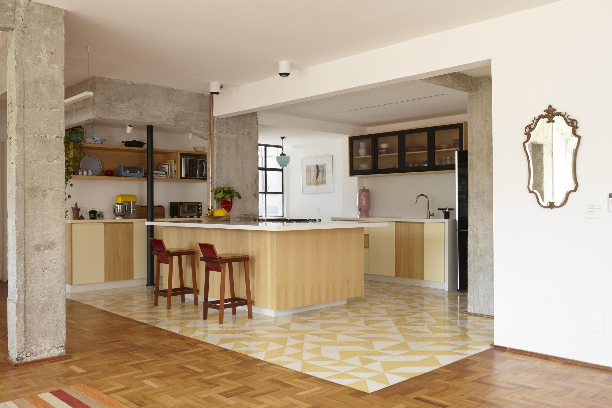 Open kitchen framed by exposed concrete columns, with a yellow patterned tile floor set into herringbone parquet and an ornate mirror