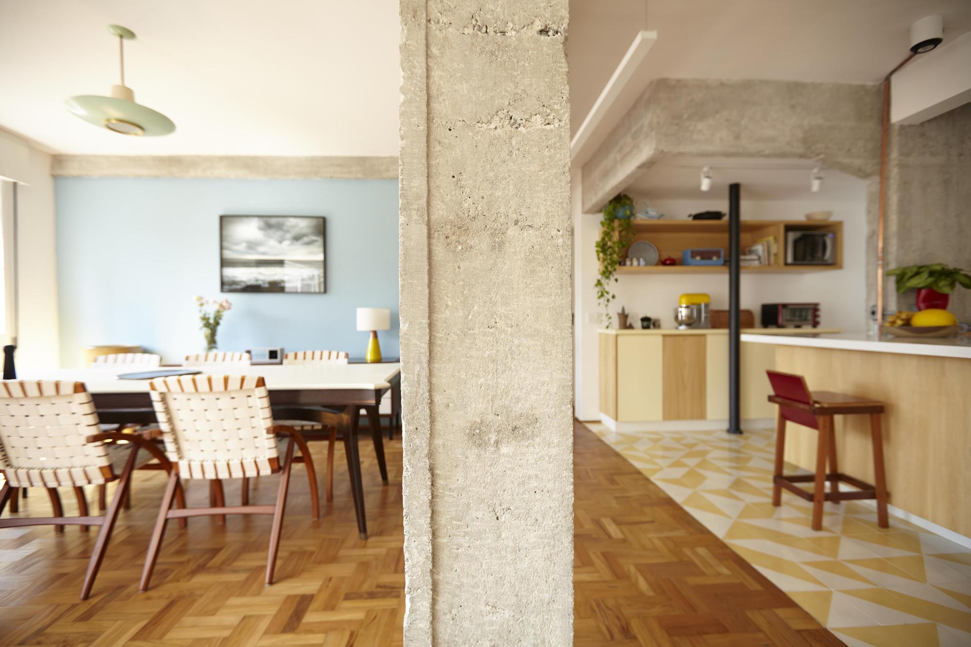 Rough concrete column dividing the dining area, with woven-strap chairs and a blue wall, from the kitchen and its tiled floor