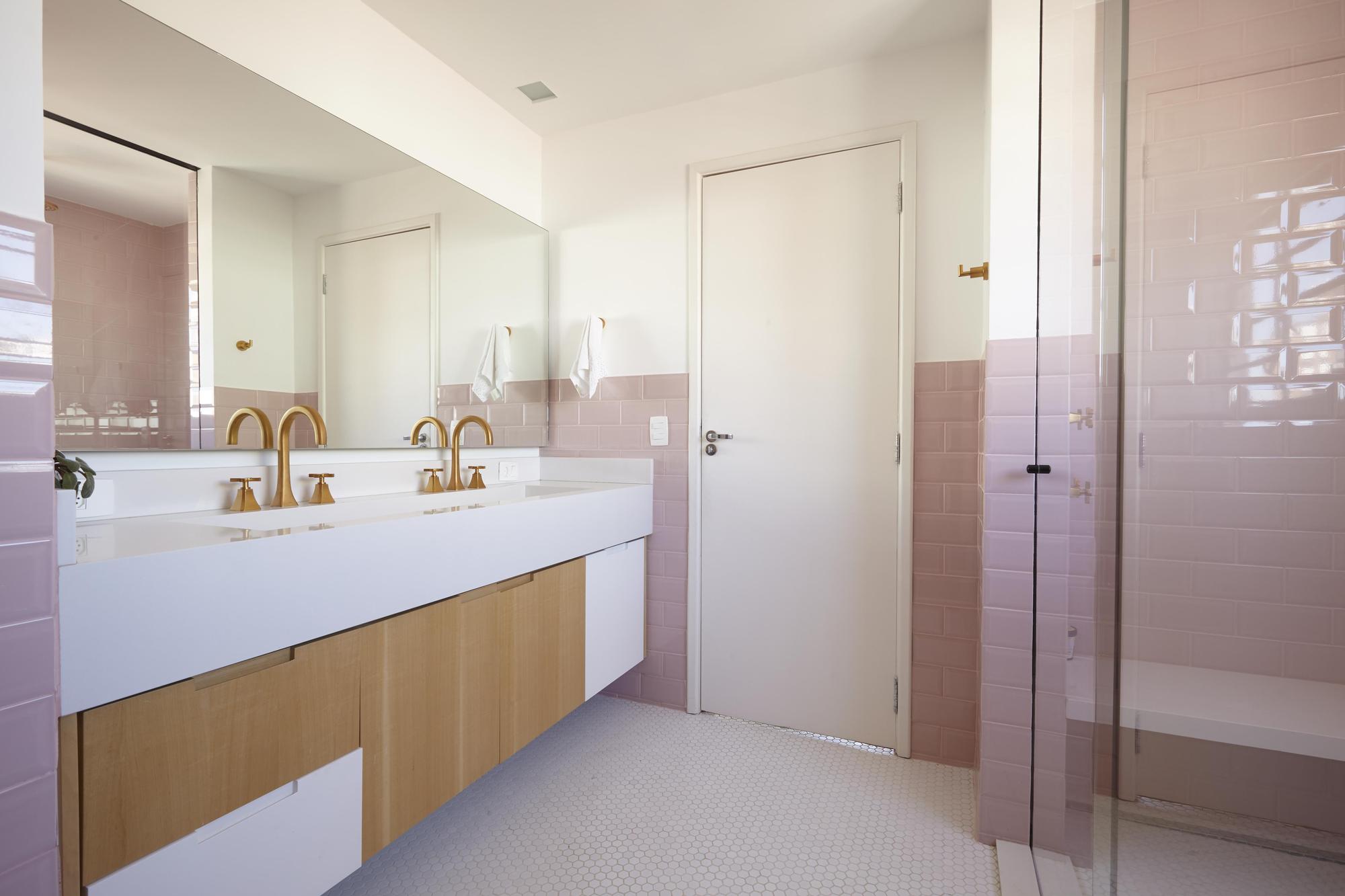Bathroom double vanity with brass taps and light wood drawers beside a white door and a pink tiled shower in soft daylight