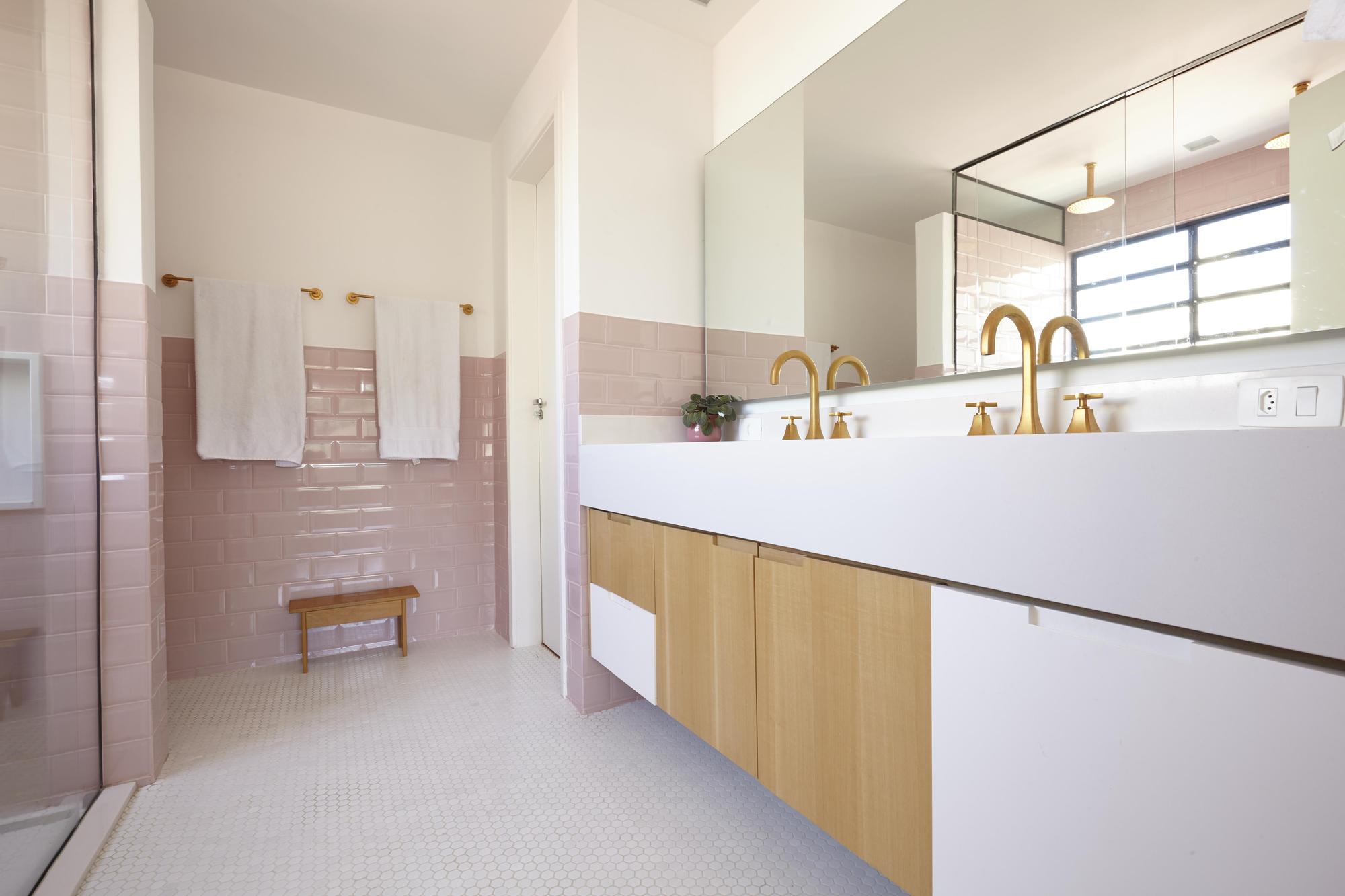 Floating double vanity with brass faucets facing a pink tiled wall with towels and a small wooden stool on hexagonal white floor tile