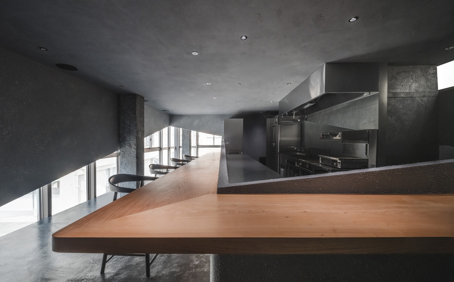 Angled timber counter wrapping a dark open kitchen, with stools lined along a band of daylight from the window wall
