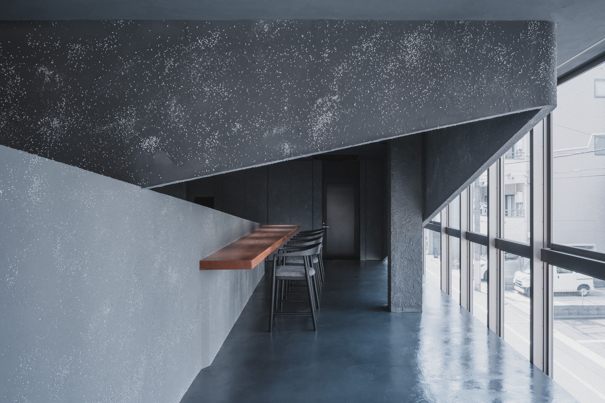 Speckled dark plaster wall and slanted ceiling above a wall-mounted wood shelf and stools, with parked cars visible through tall windows