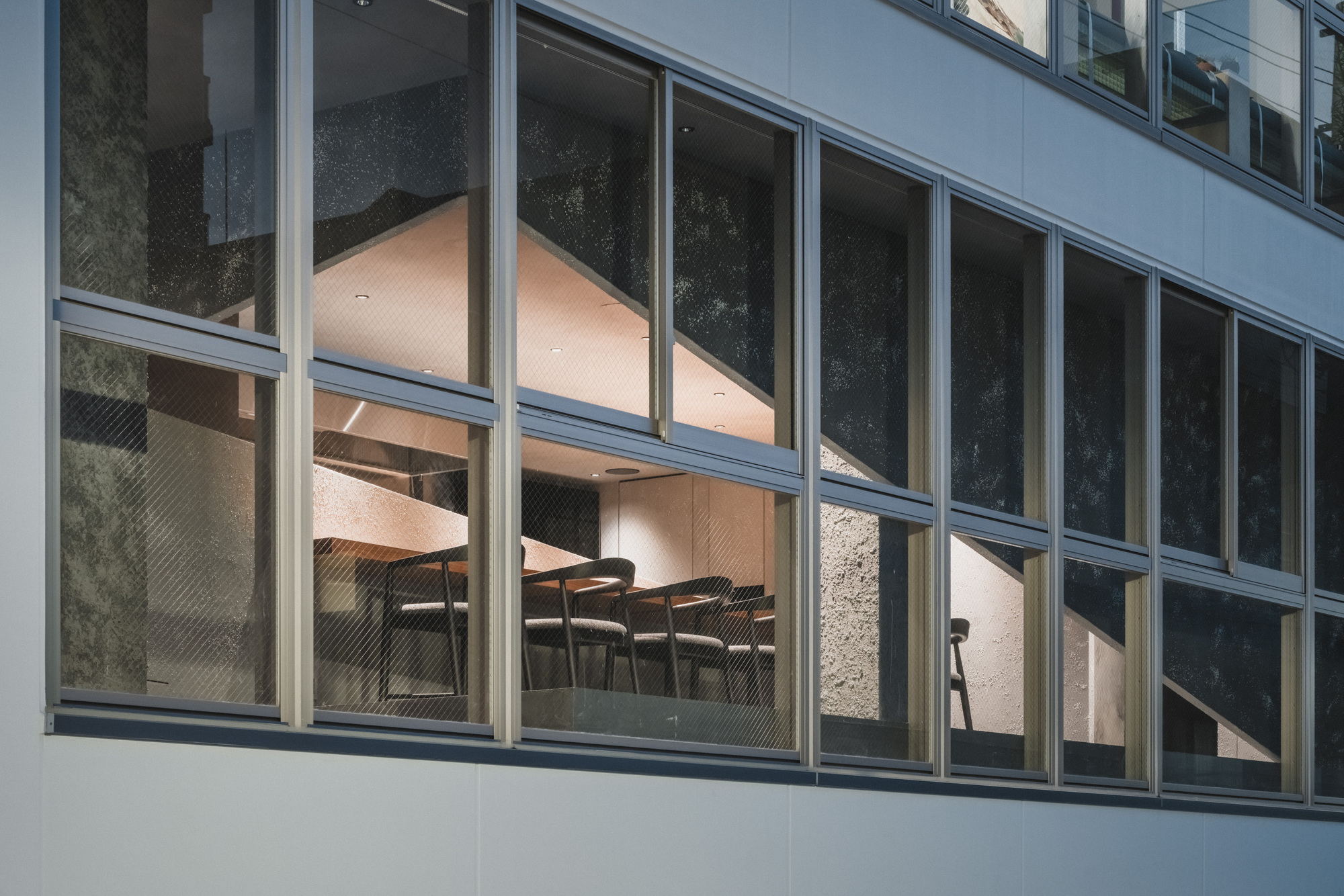 Exterior view of the gridded window facade, with stools and a textured dark interior wall glowing at dusk