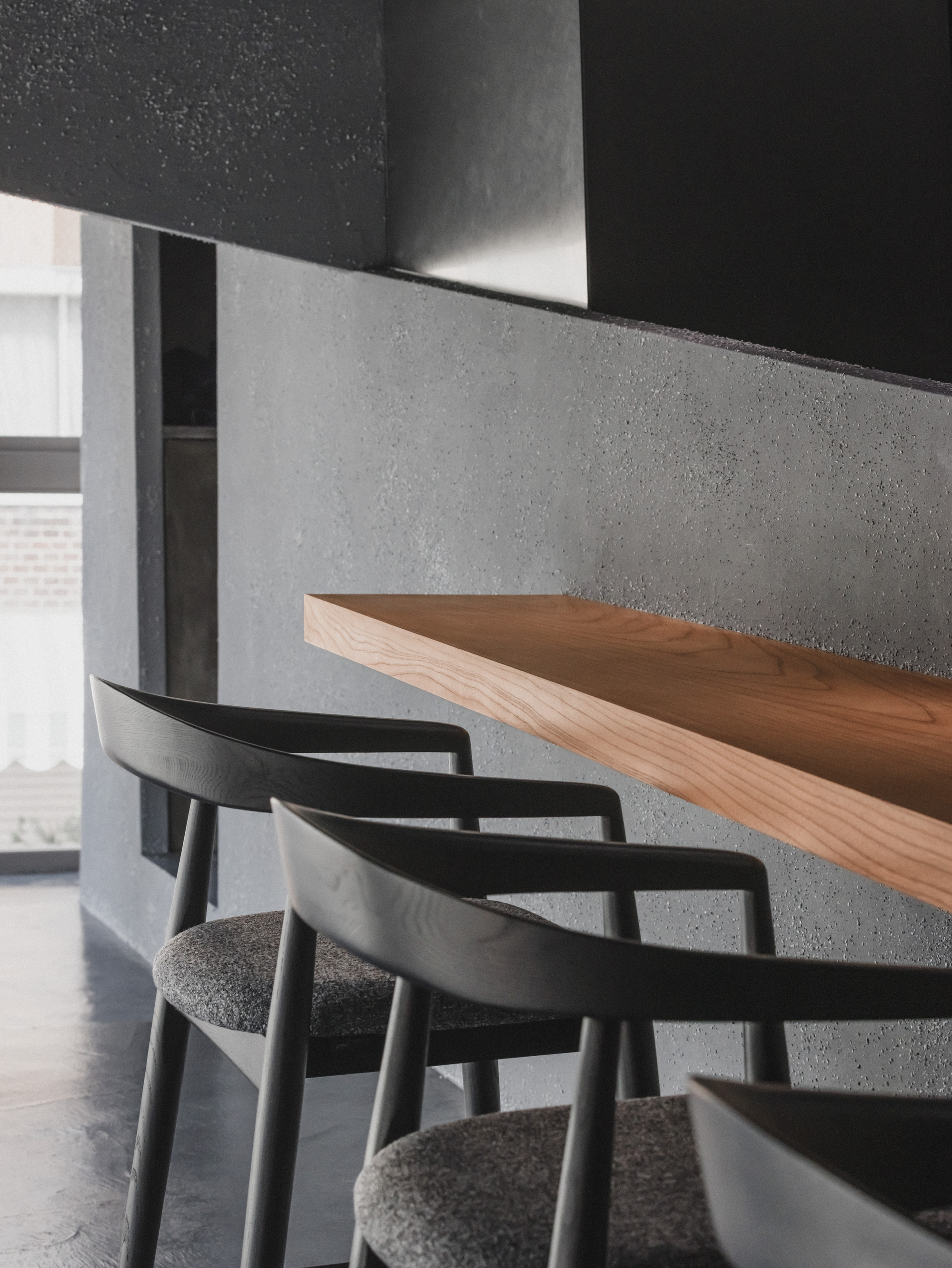 Close-up of black wooden armchairs with grey upholstered seats beside a cantilevered timber shelf on a rough grey wall