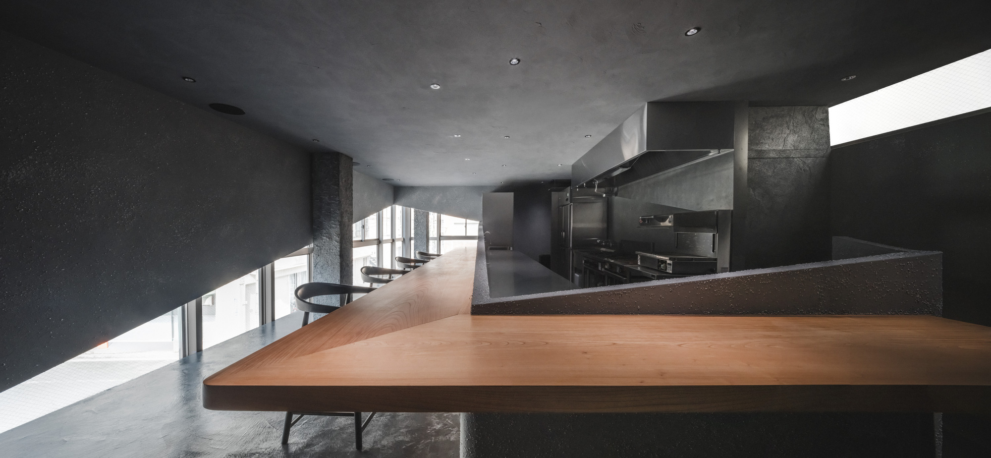 Wide view of the long timber counter angling toward the steel kitchen, with a bright window slot at right