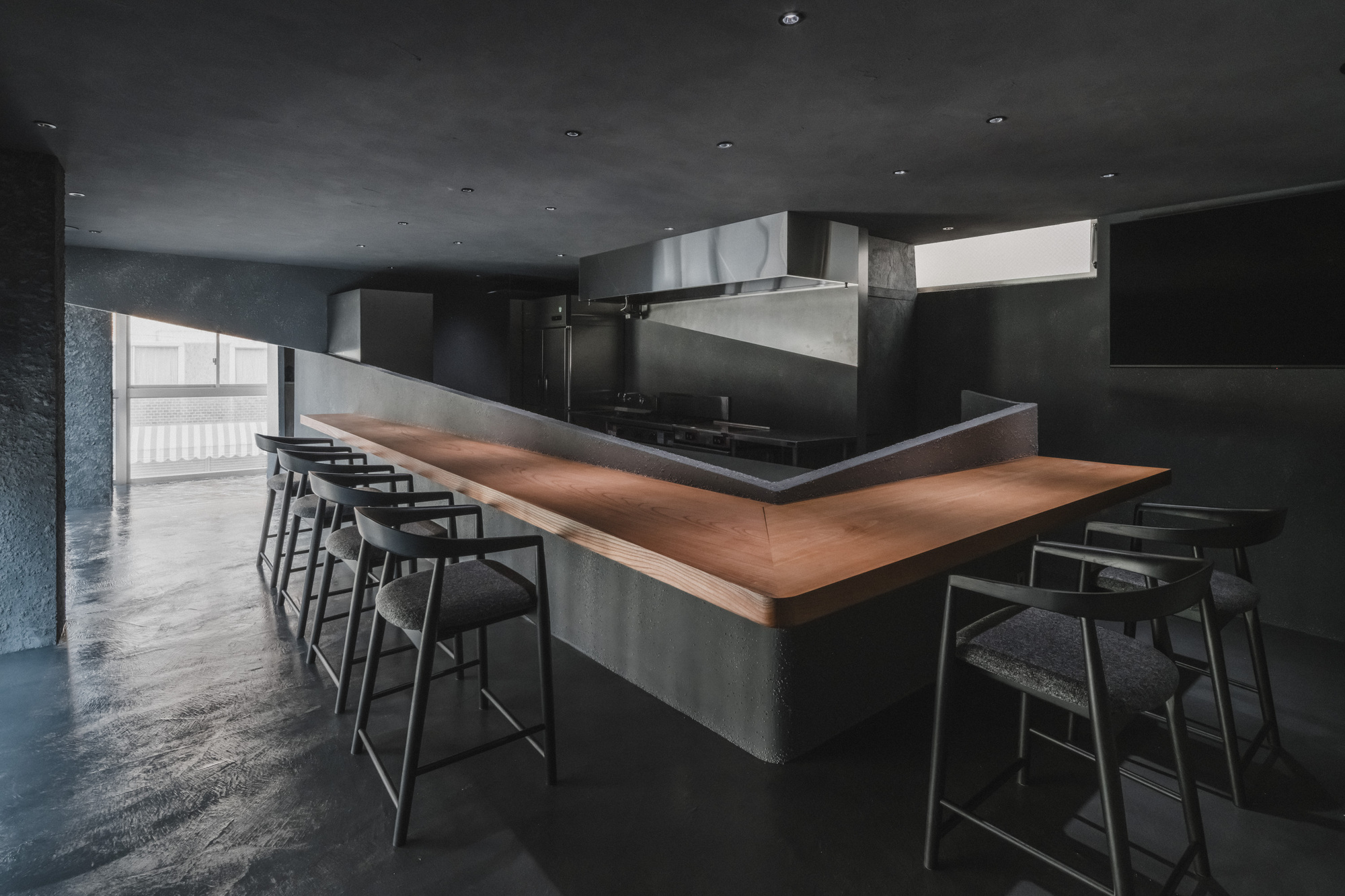 V-shaped timber counter wrapped in rough grey plaster with upholstered stools facing an open kitchen, daylight spilling from a high window