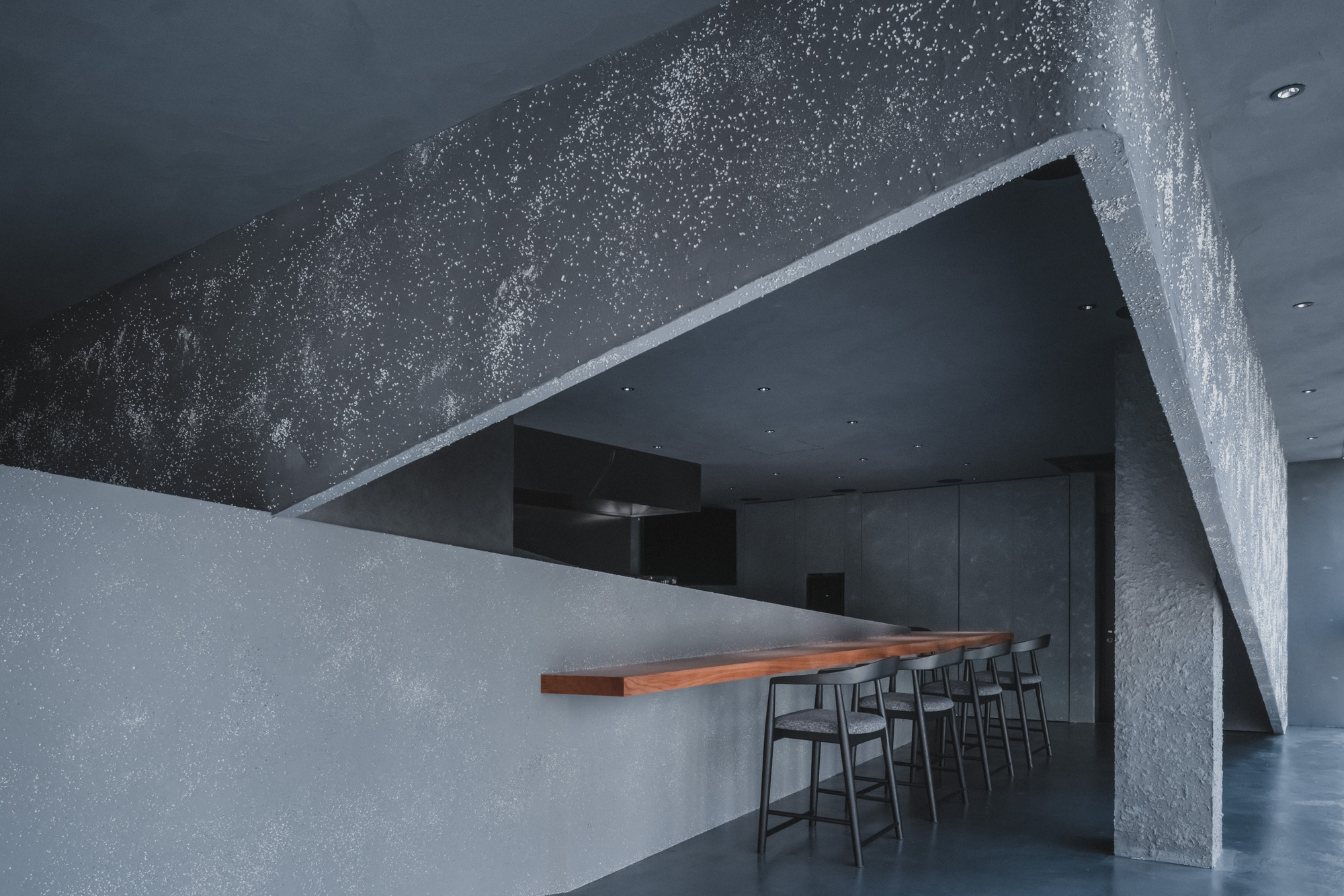 Angular plaster soffit flecked with white aggregate above a cantilevered wood counter and a row of stools in cool light