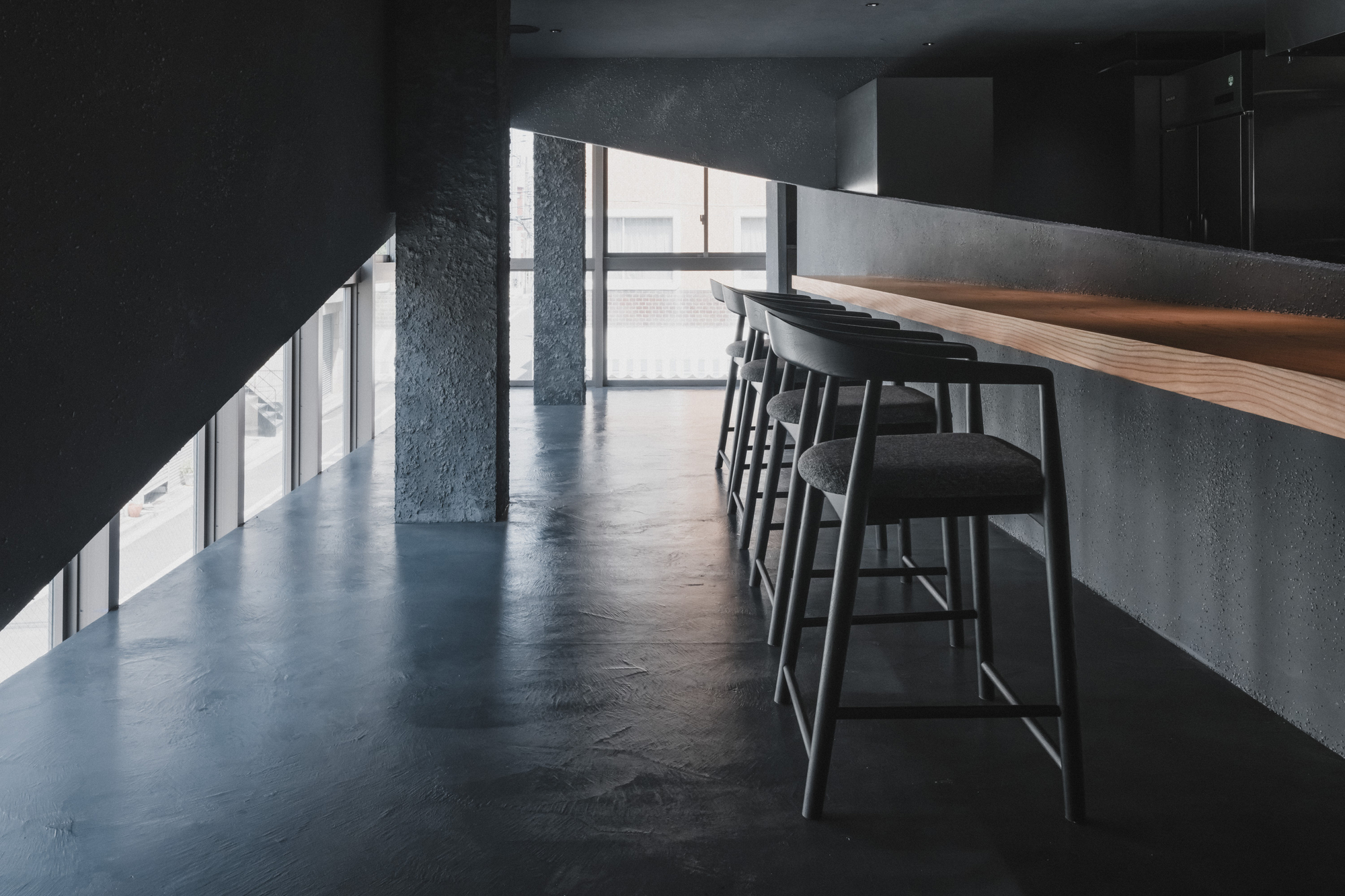 Row of black stools along the plaster counter beside a rough textured column and a slanted glass wall in afternoon light