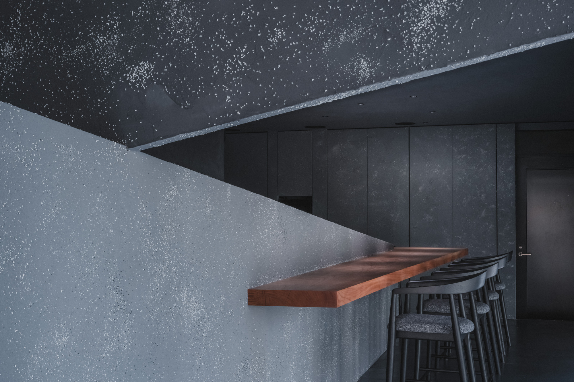 Close view of the speckled plaster ceiling edge and wall above a floating red-brown wood counter with grey upholstered stools